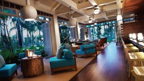 A spacious lounge area with wooden flooring, blue seating, and large windows offering views of tropical foliage.