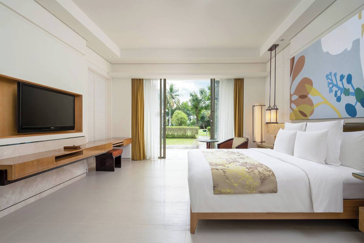 A modern hotel room with a large bed, a flat-screen TV, a wooden console table, and a view of the garden through a glass door.