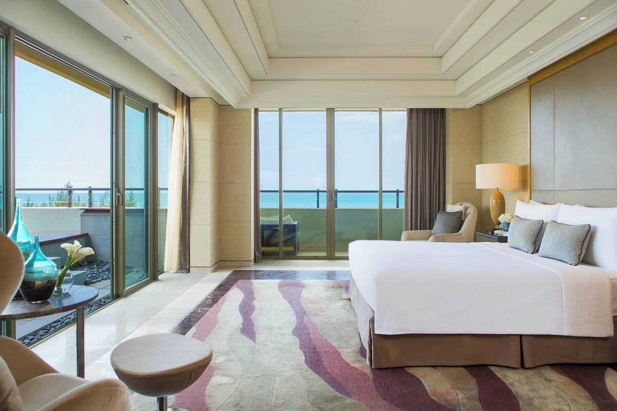 A hotel room with a large bed, a sitting area with a chair and table, a view of the sea through large windows, and a balcony.