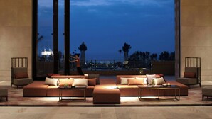 A modern lounge area with a large sofa set, a coffee table, and two armchairs. A large window provides a view of the cityscape at dusk.