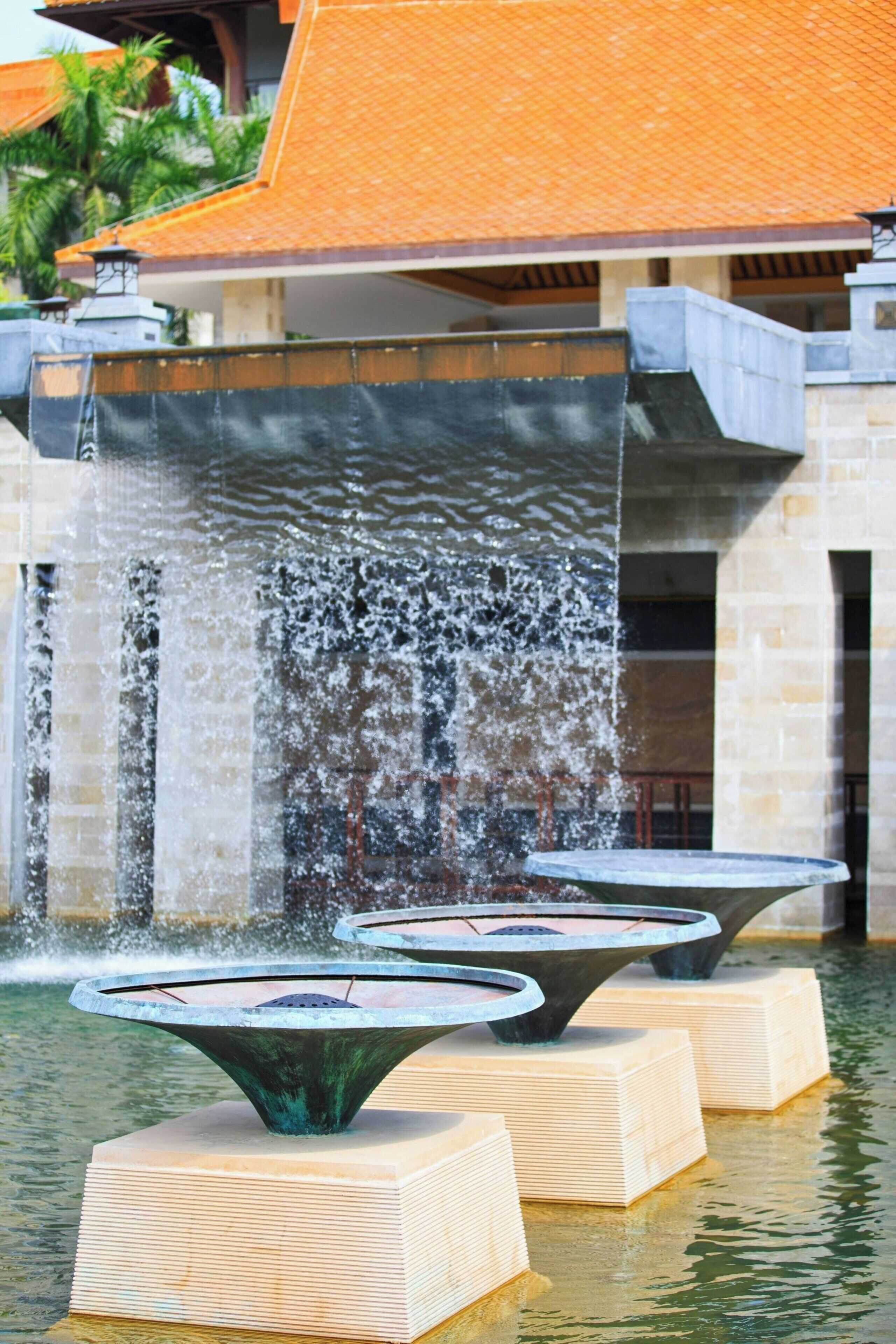 A fountain with multiple tiers of water jets in front of a building with a tiled roof.
