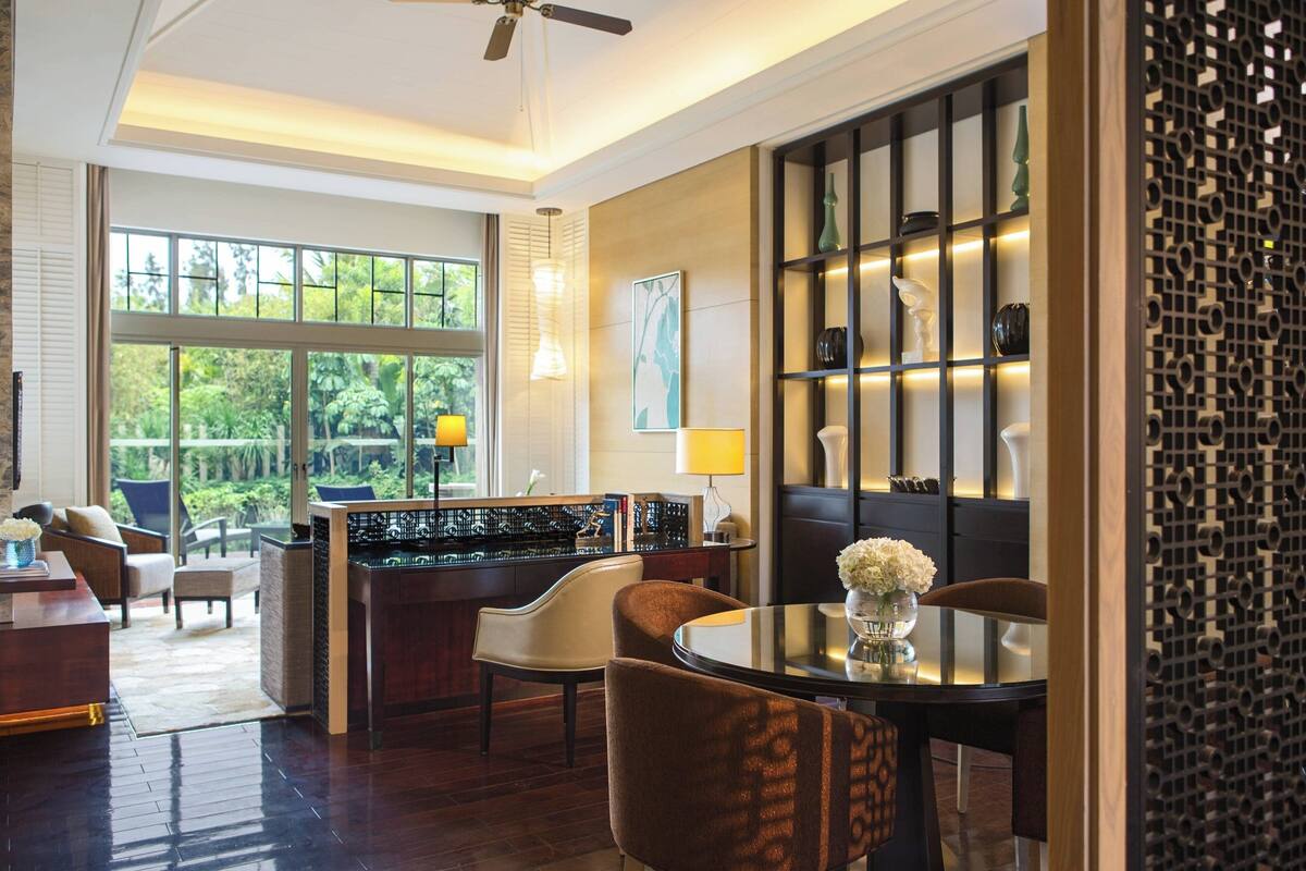 A modern hotel room with a dining area, a bar, and large windows offering a view of greenery.