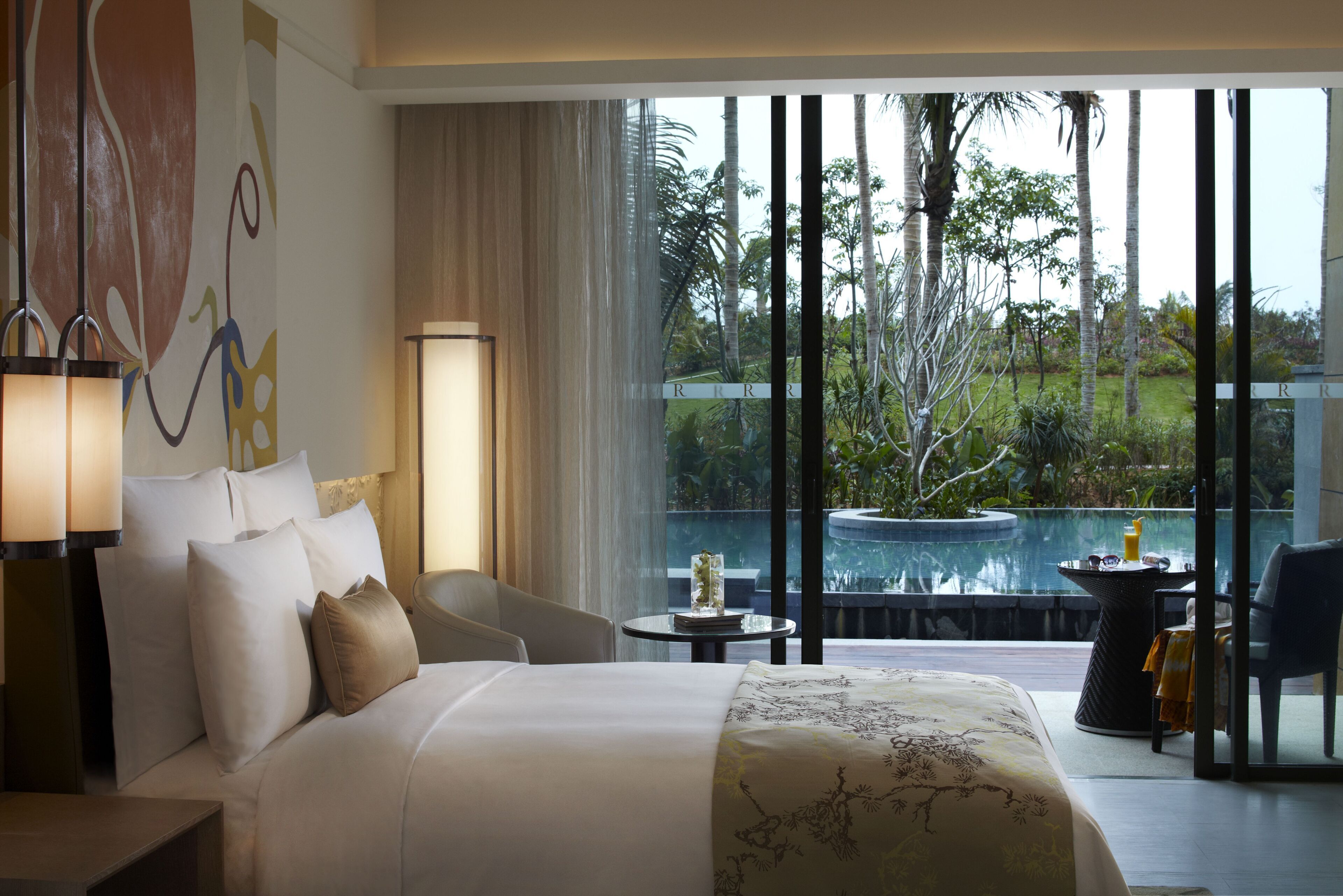 A hotel room with a large bed, a view of a swimming pool, and a balcony with a table and chairs.
