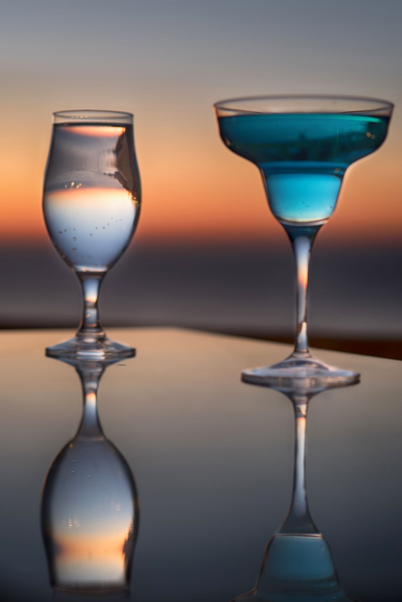 Two glasses on a reflective surface with a sunset in the background.