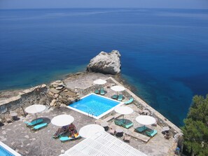 Uma área de piscina com espreguiçadeiras e guarda-sóis, com vista para um mar azul e límpido.