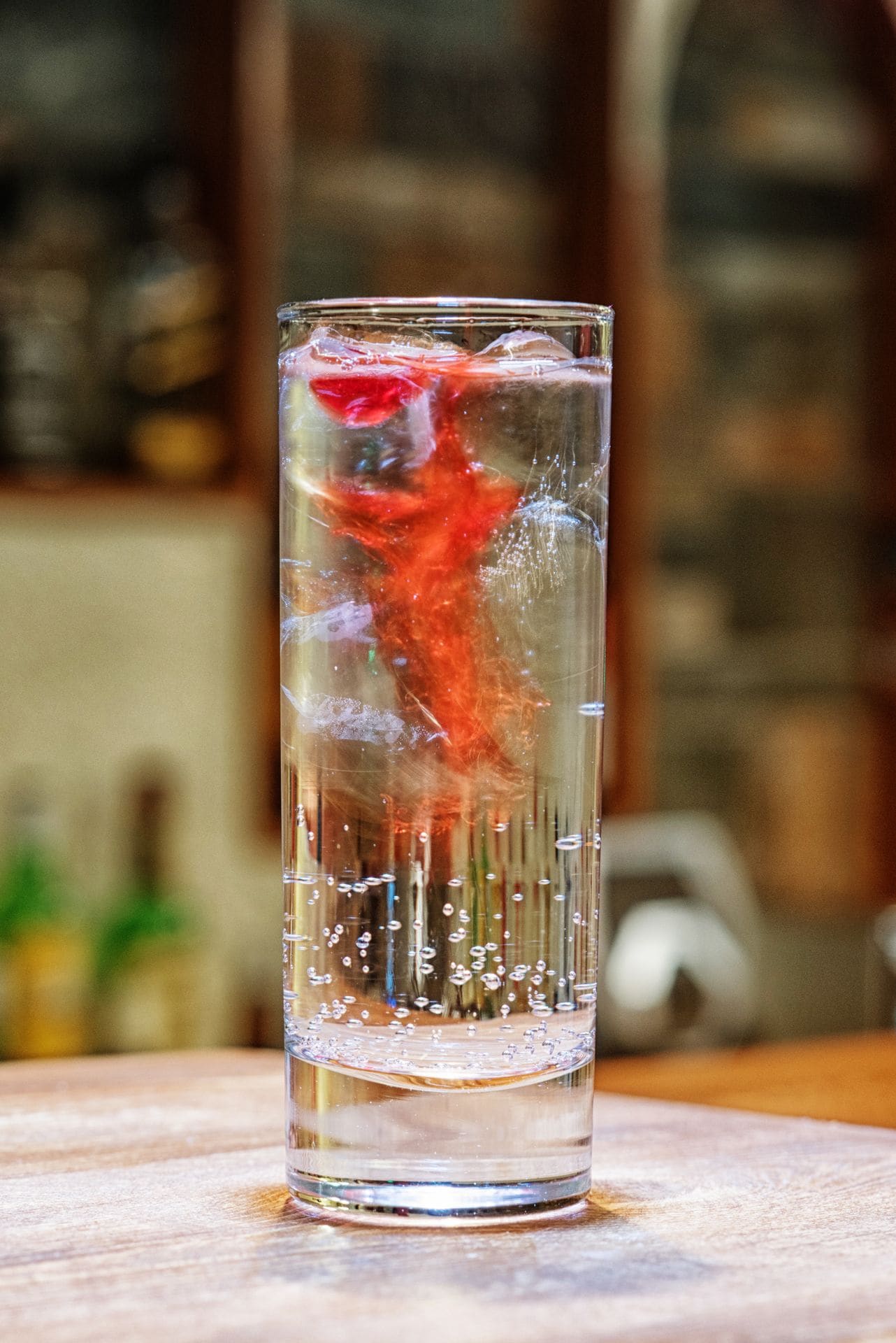 Un vaso transparente con cubitos de hielo y un líquido rojo, probablemente un cóctel, sobre una superficie de madera.