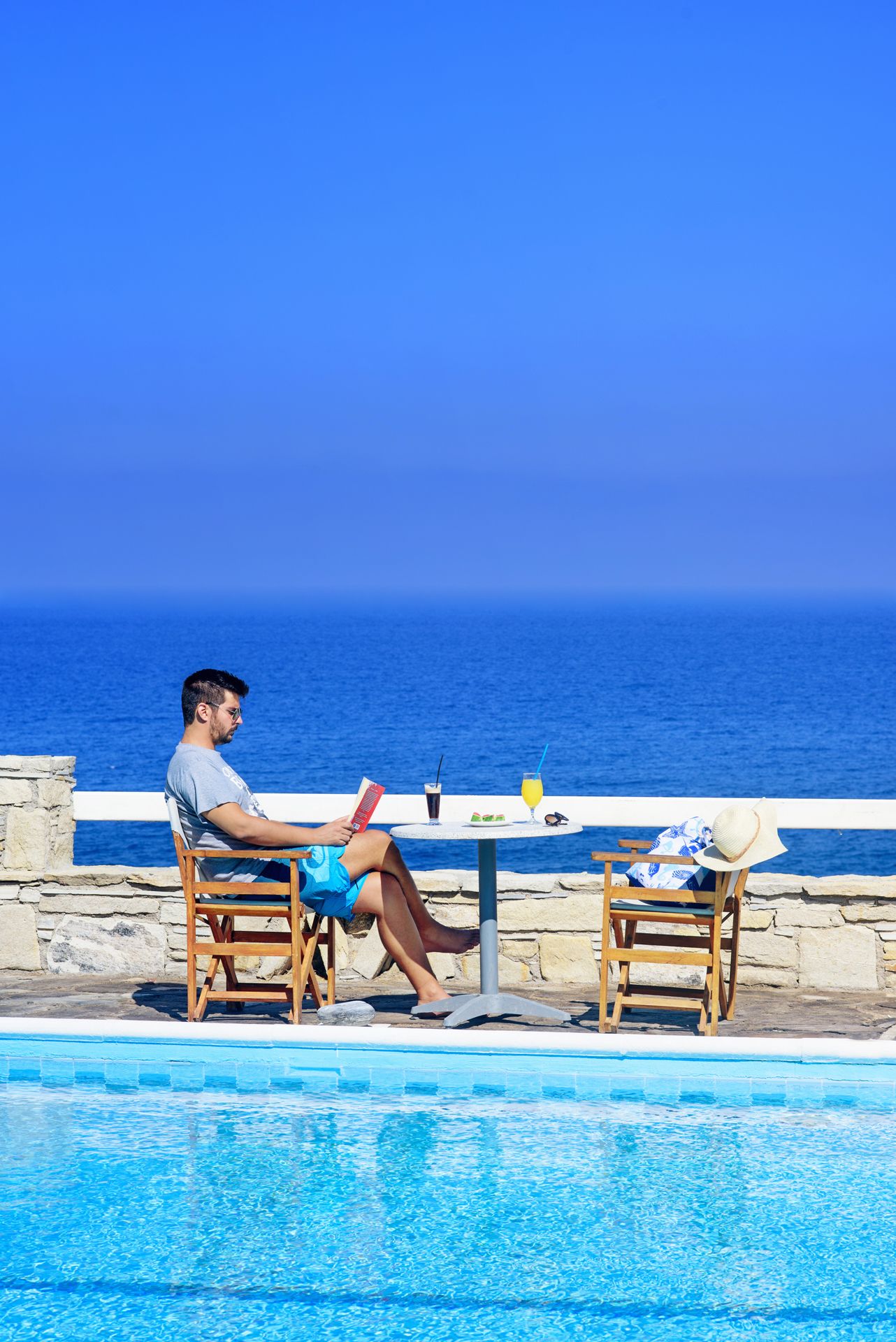 A man reading by a poolside table with a drink and a hat, overlooking a clear blue sea.