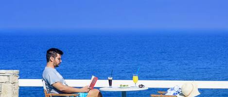 A man reading by a poolside table with a drink and a hat, overlooking a clear blue sea.