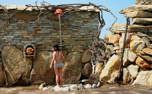 A person in a bikini standing in front of a stone wall with a water spout.