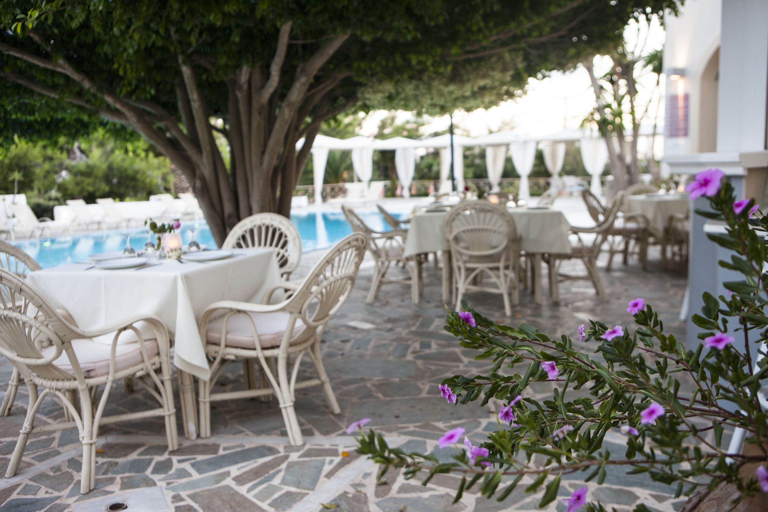 An outdoor dining area with white wicker furniture and a stone floor, surrounded by greenery and a pool.