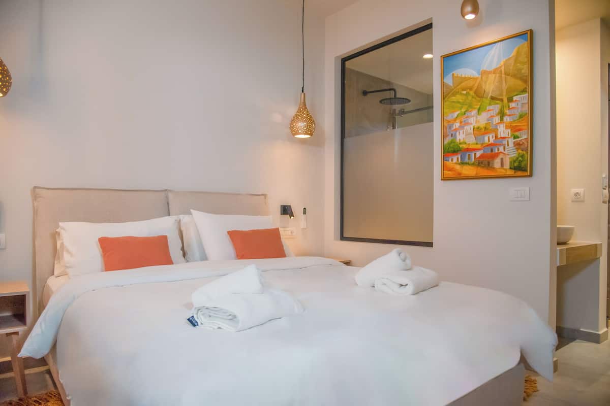A hotel room with a large bed, white bedding, orange pillows, a painting on the wall, and a shower area.