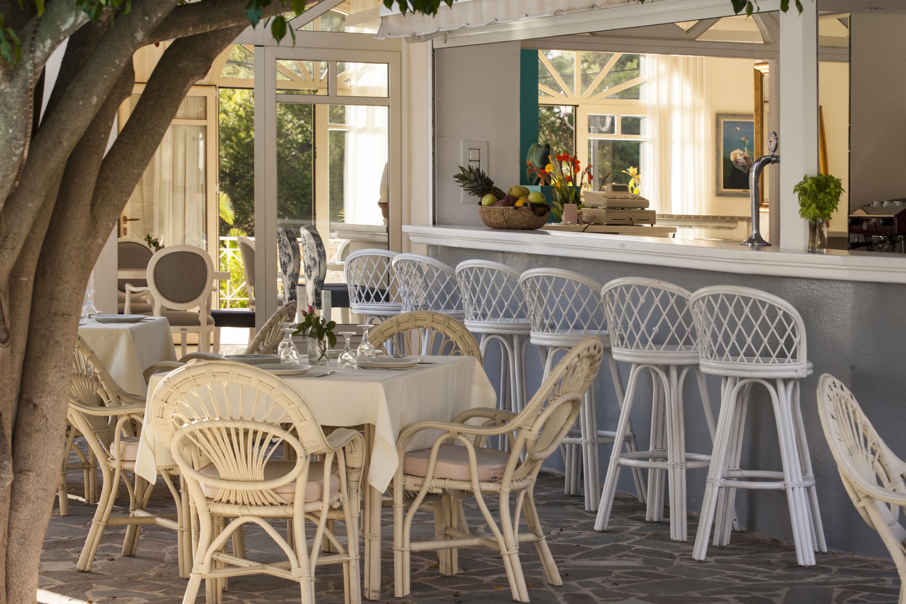 Een terras met een eethoek, rieten stoelen en tafels, een bar met een fruitmand, en uitzicht op het interieur door een glazen deur.