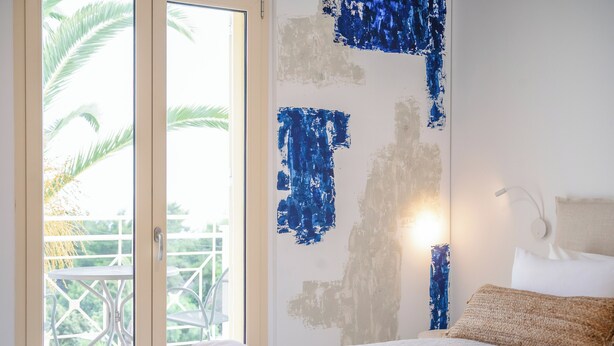 A hotel room with a bed, a nightstand, a lamp, and a view of a balcony with a palm tree. - Princess Hotel (Kefalonia)