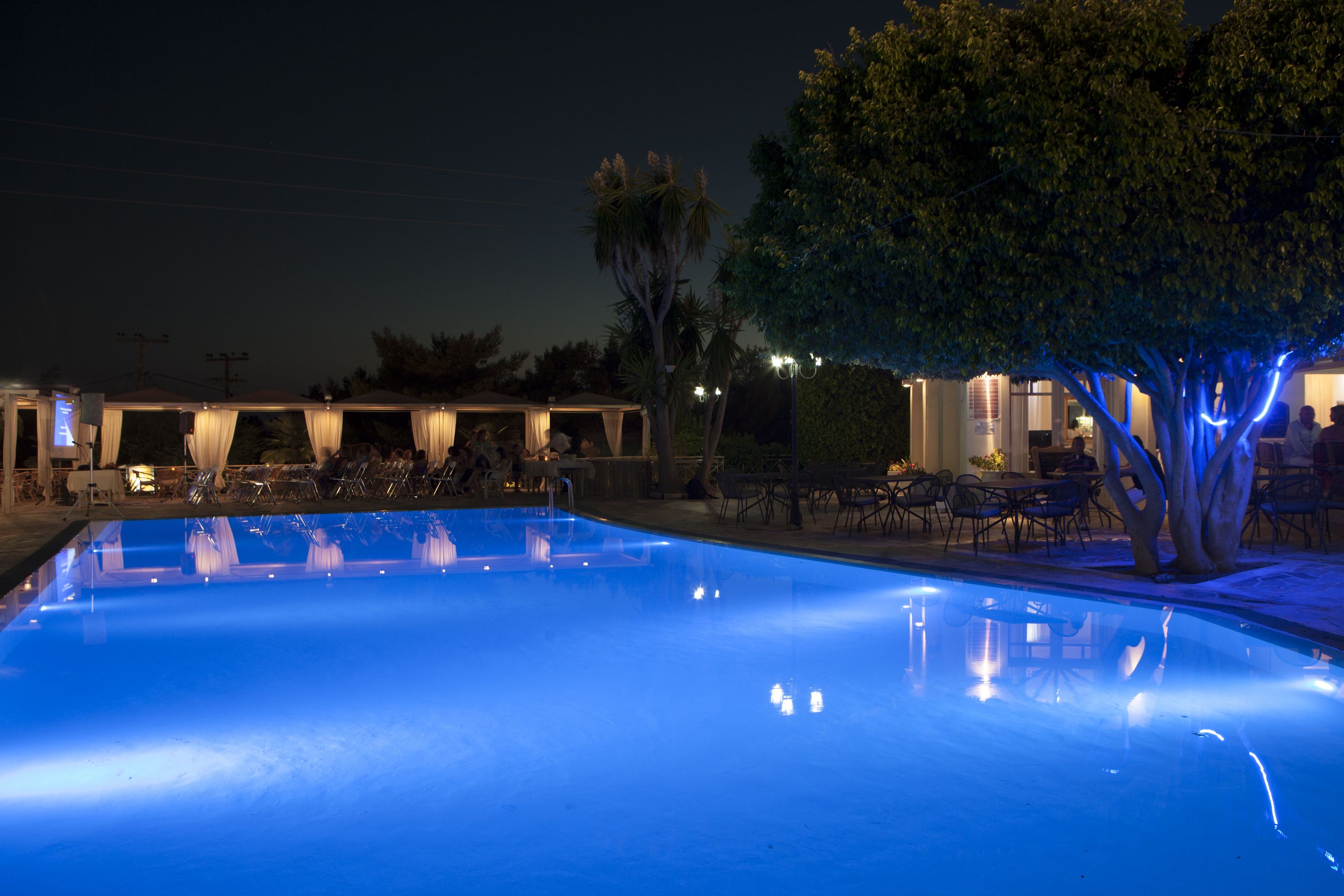 A swimming pool with a blue-lit edge, surrounded by outdoor seating and a building with a covered patio.