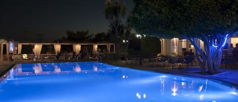 A swimming pool with a blue-lit edge, surrounded by outdoor seating and a building with a covered patio.