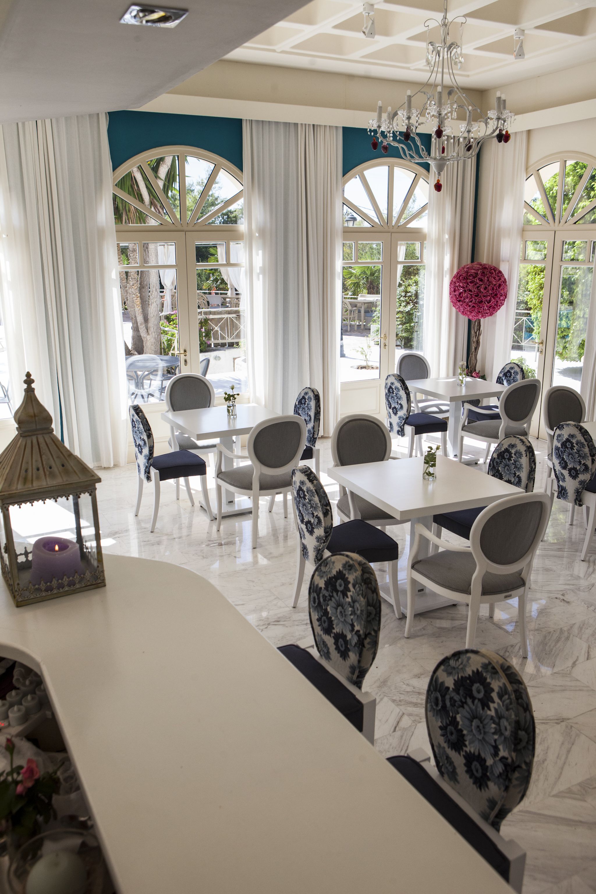 Un espace repas avec des tables et des chaises blanches, de grandes fenêtres avec des rideaux et un lustre.