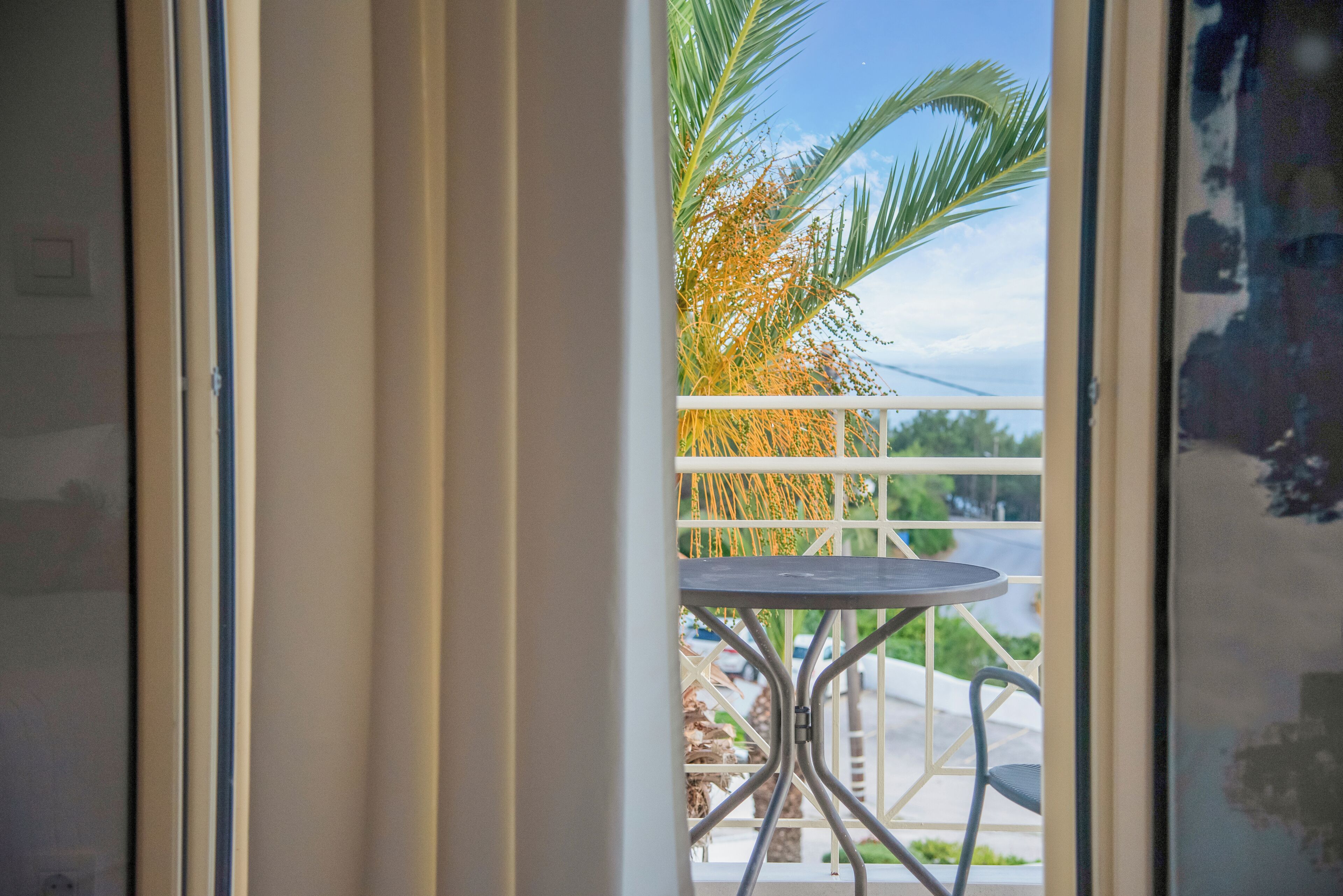 A view from a balcony with a metal table and chairs, a palm tree, and a street in the background.