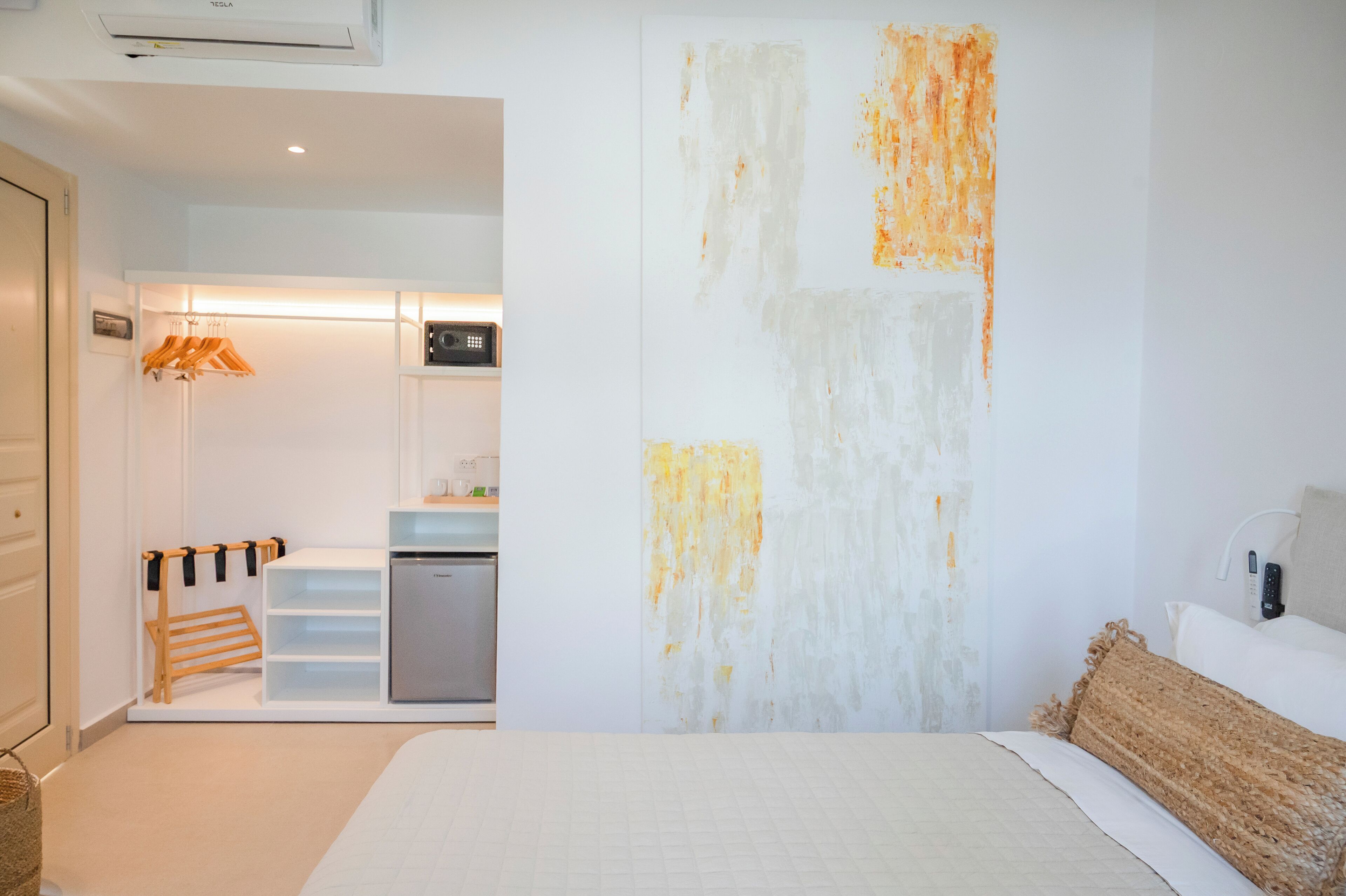 A modern hotel room with a bed, a bedside table, a wall art piece, and a kitchenette area.