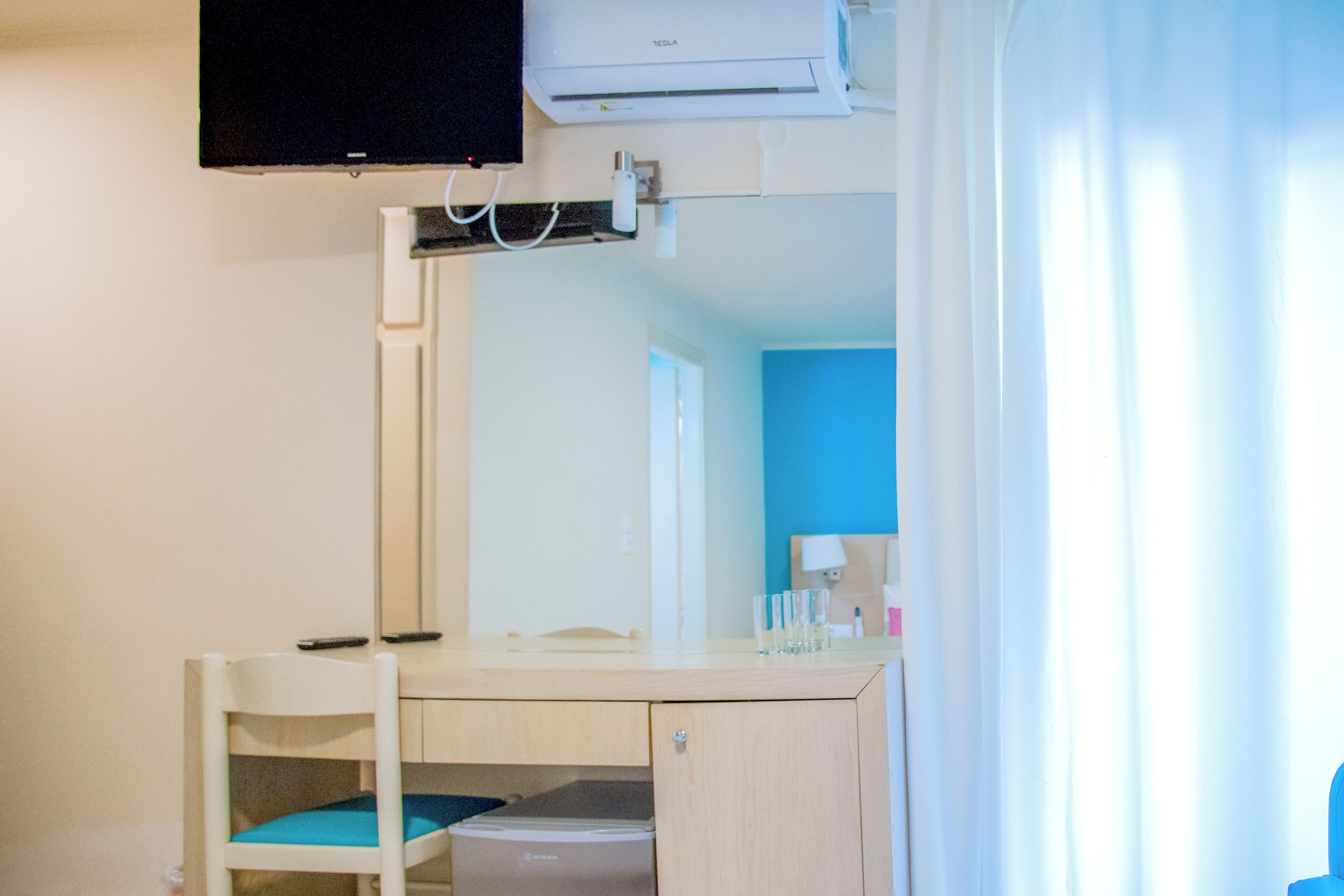 A room with a bed, a desk, and a chair. There is a mirror above the desk and a window with curtains.