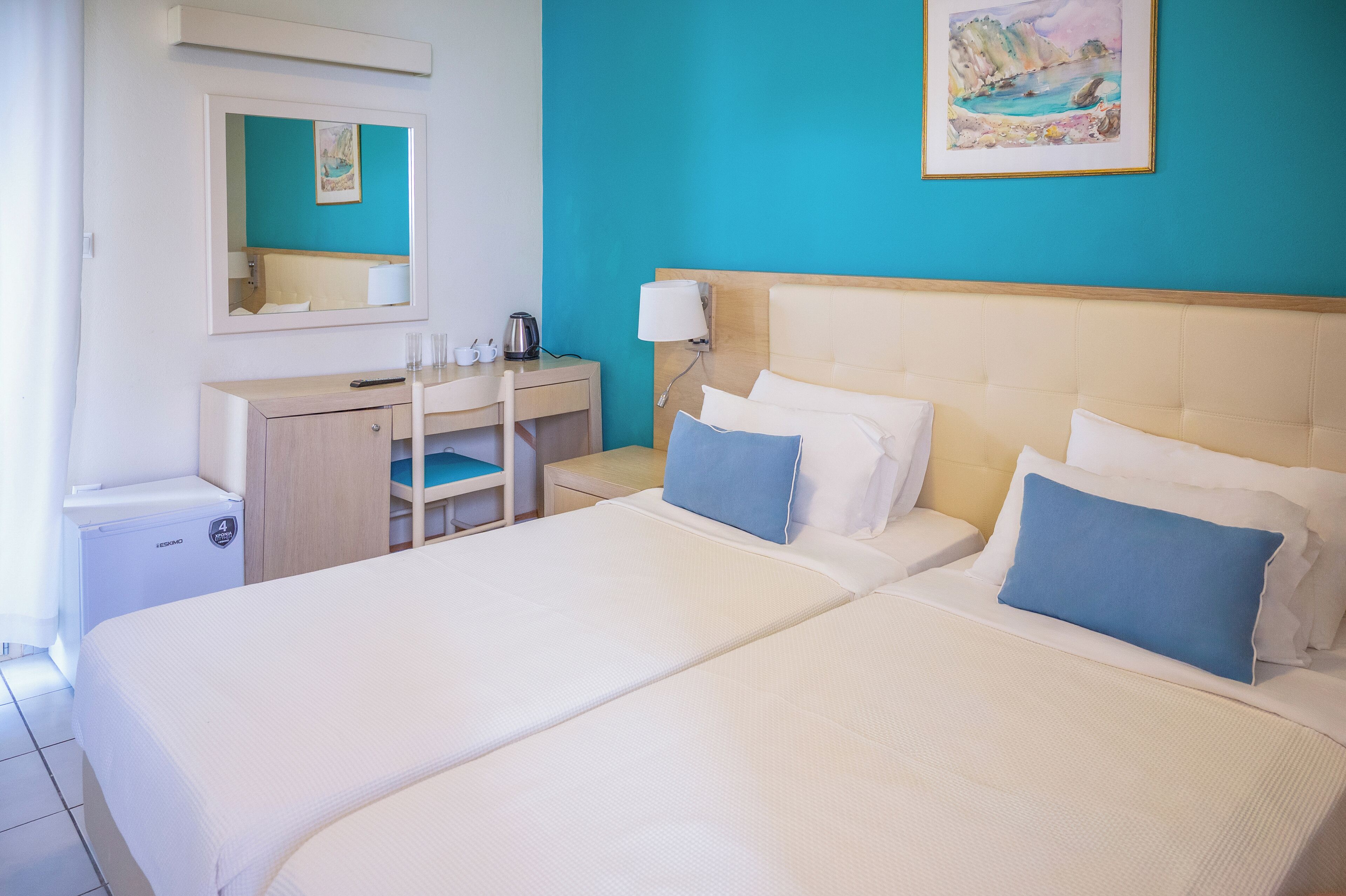 A hotel room with two beds, a desk, a chair, a small table, a mirror, and a painting on the wall.