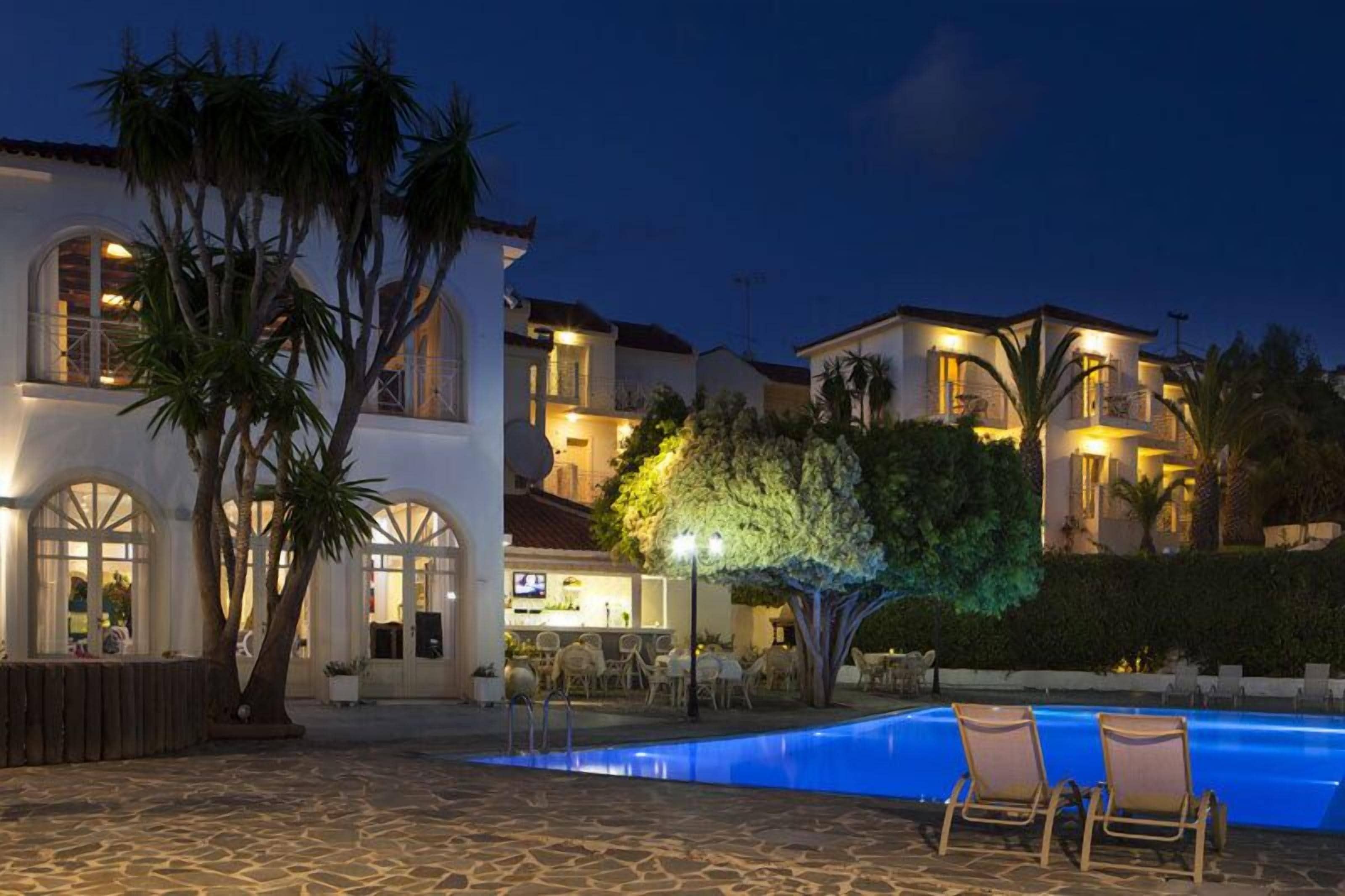 A poolside area with a stone floor, lounge chairs, and a well-lit building with multiple windows and a bar area.