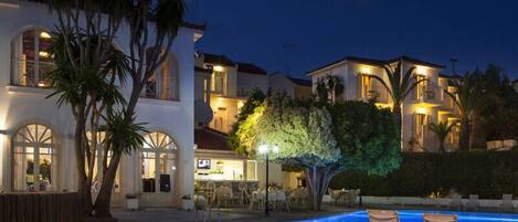 A poolside area with a stone floor, lounge chairs, and a well-lit building with multiple windows and a bar area.