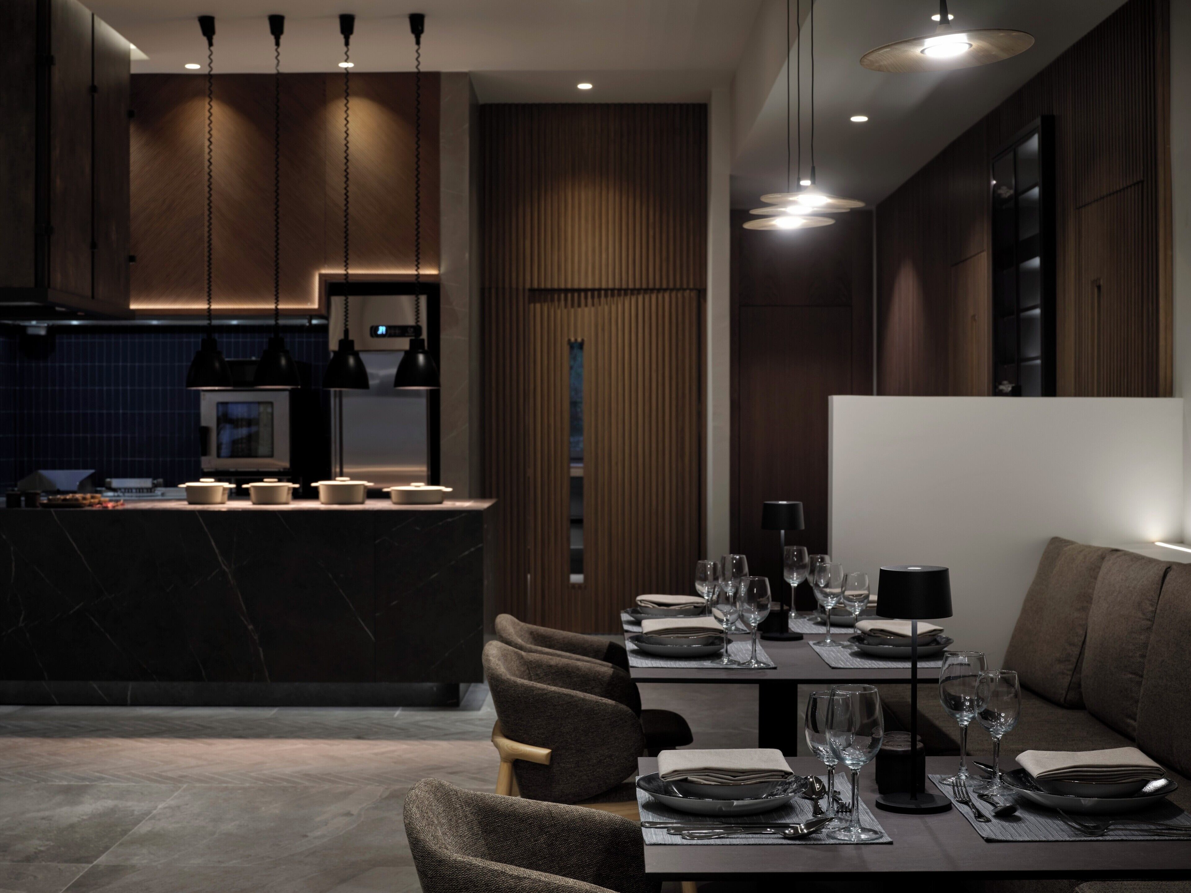 Un espace repas moderne avec une table aux tons foncés, dressée pour deux personnes, un coin bar doté d’un comptoir en marbre et un éclairage suspendu.