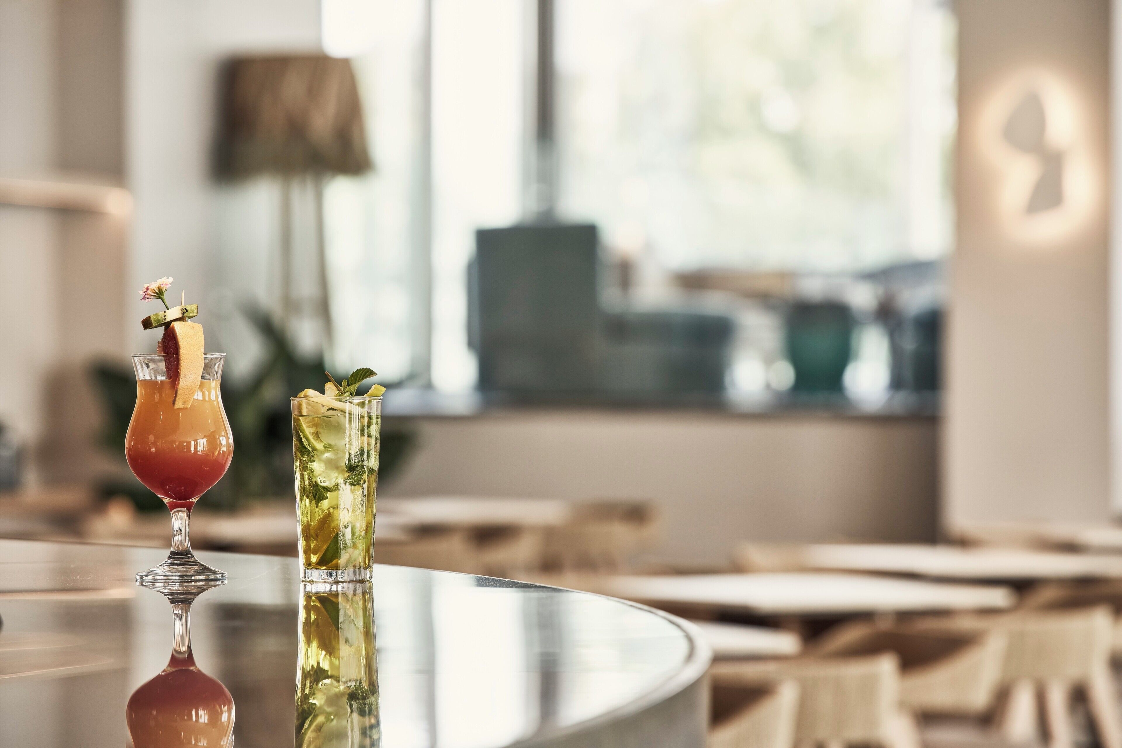 A bar area with a glass of red drink and a glass of iced beverage, a lamp, and a view of the outdoors.