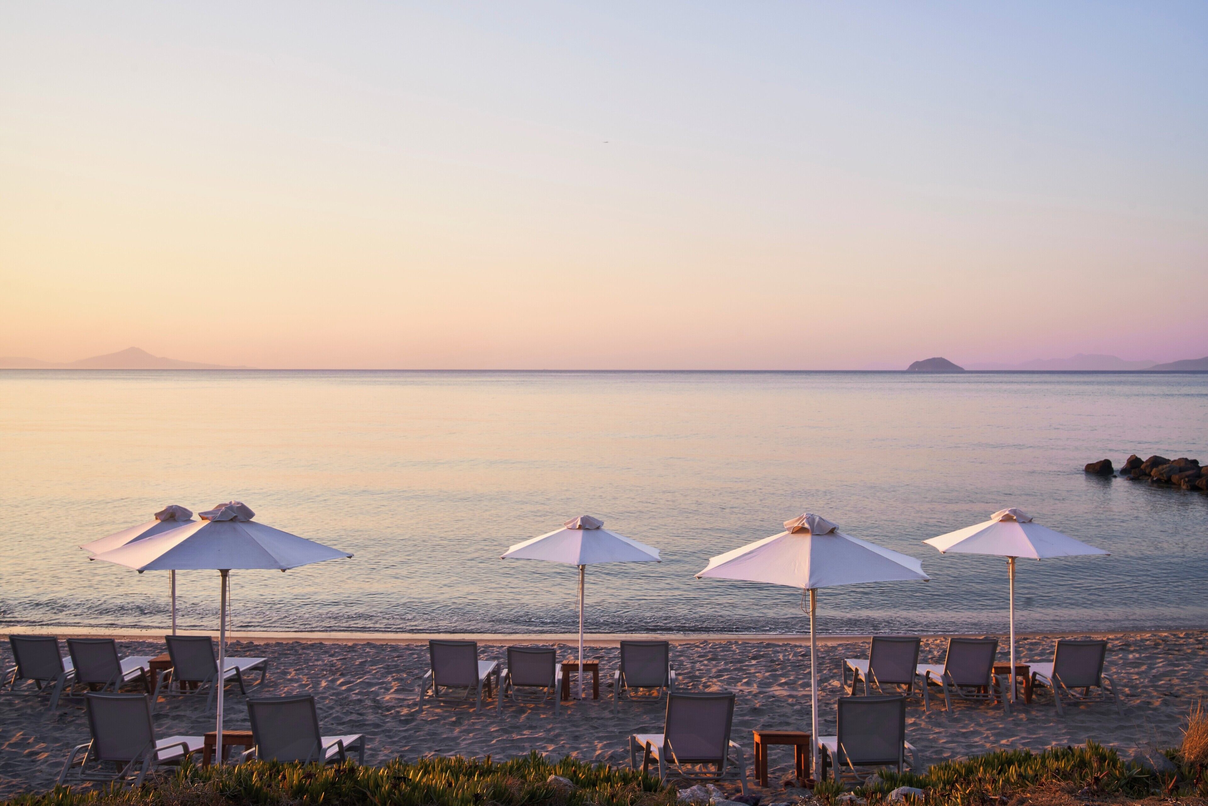 Una playa con sillas de playa y sombrillas, un mar tranquilo y montañas a lo lejos.