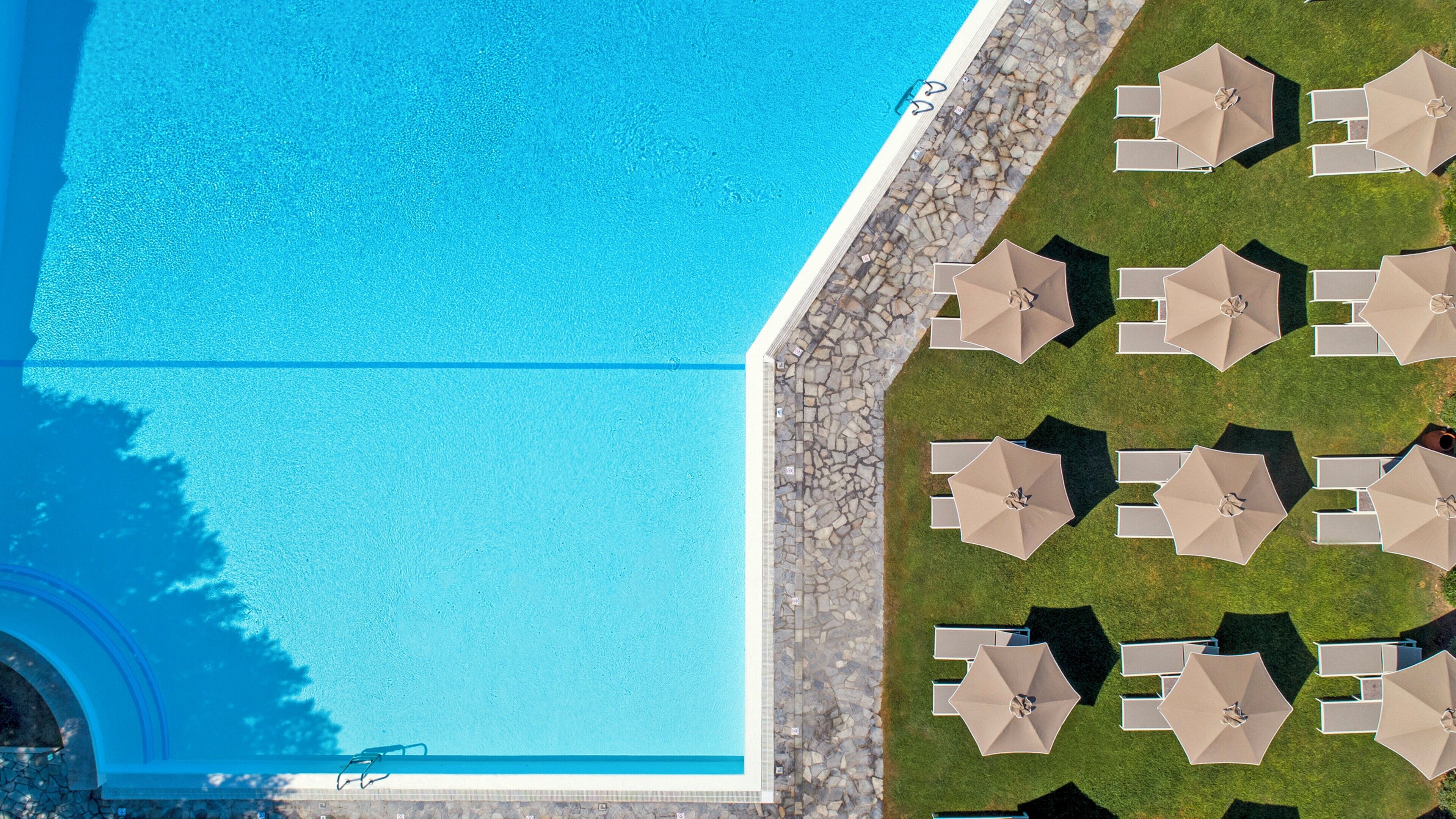 Vista aérea de una piscina con borde de piedra y múltiples sombrillas de descanso a la derecha.
