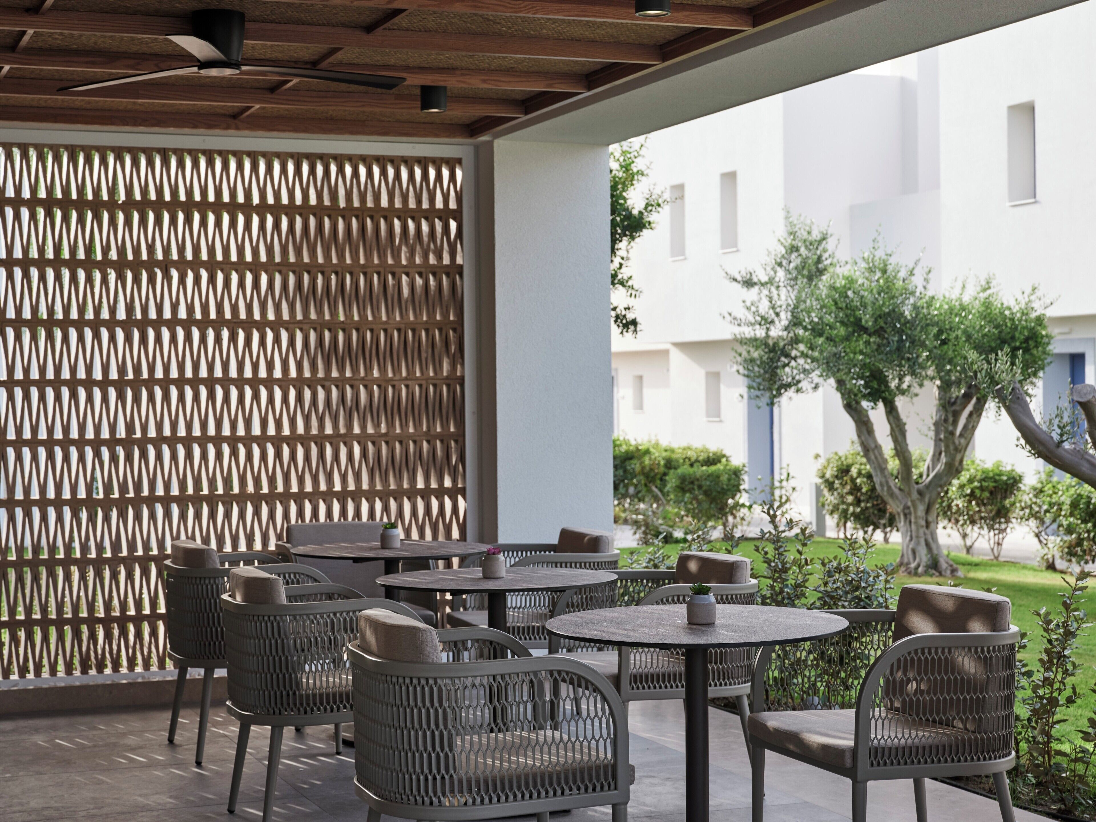 Un patio al aire libre con mesas redondas y sillas de mimbre, techo de madera y vista a un edificio blanco y vegetación.