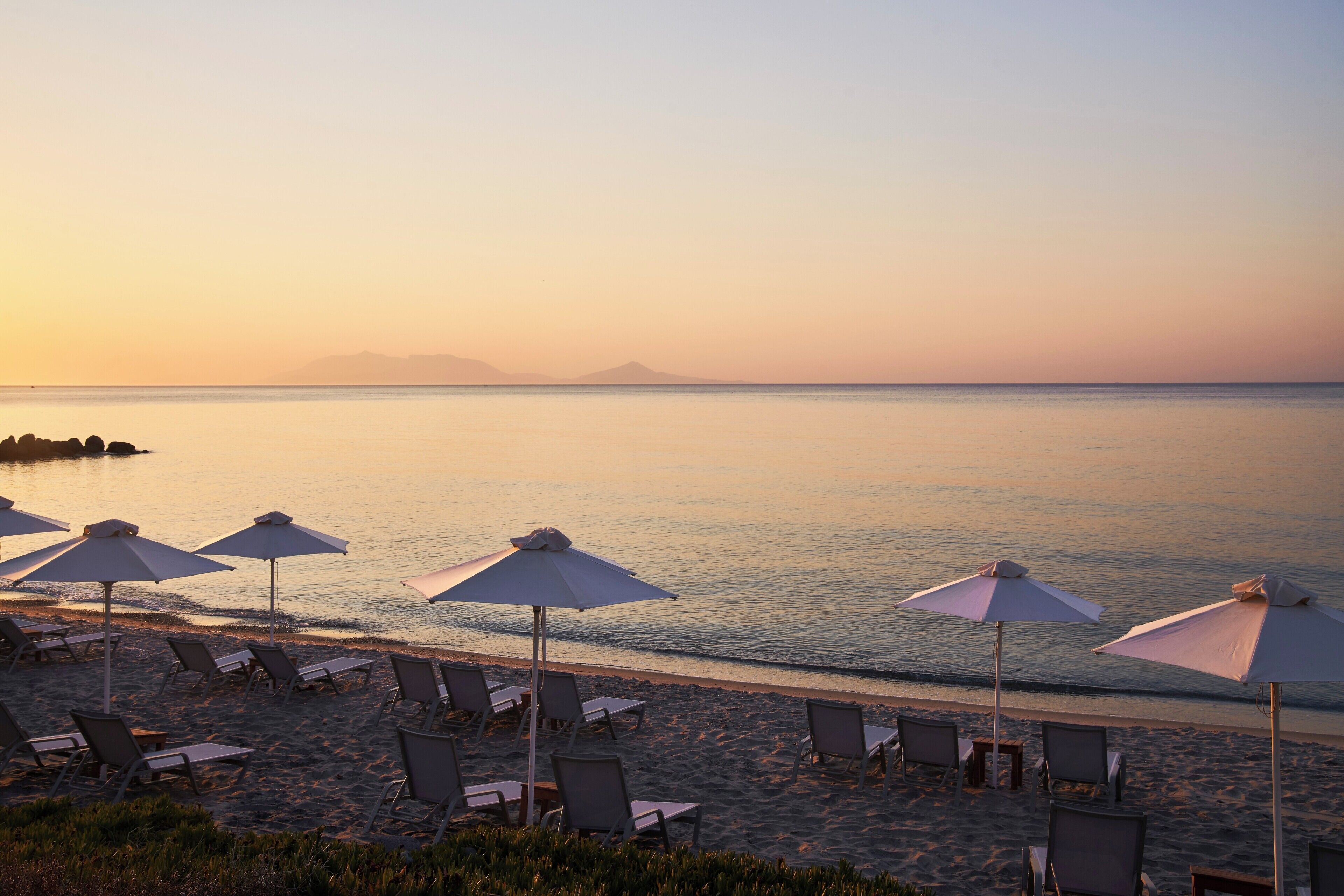 A beach with sun loungers and umbrellas, a calm sea, and a distant mountain.