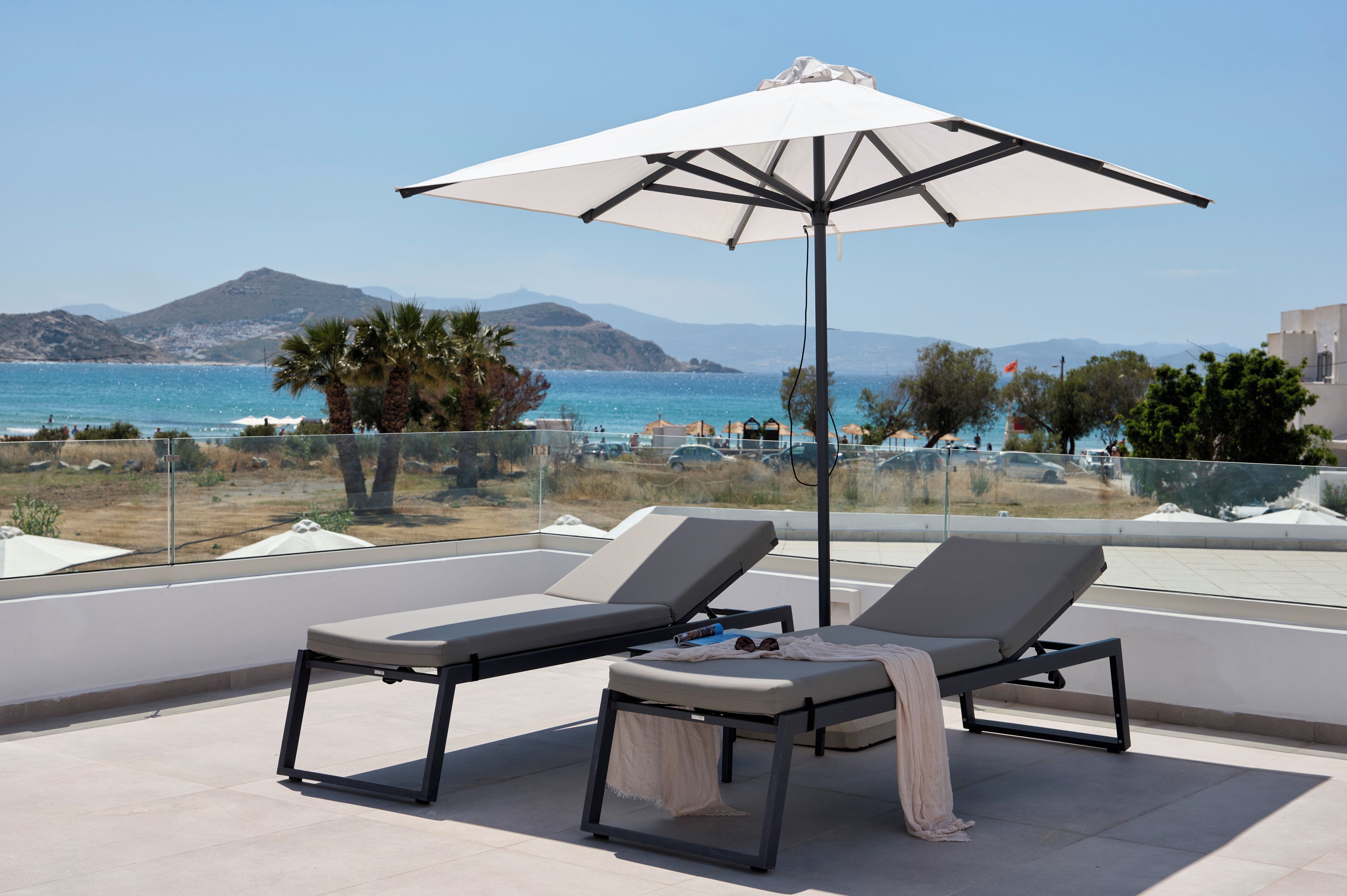 A terrace with two lounge chairs and a large umbrella, overlooking a beach and the sea.