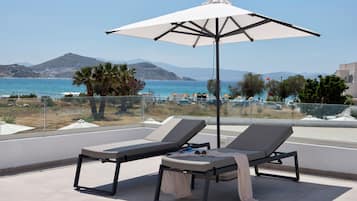 A terrace with two lounge chairs and a large umbrella, overlooking a beach and the sea.