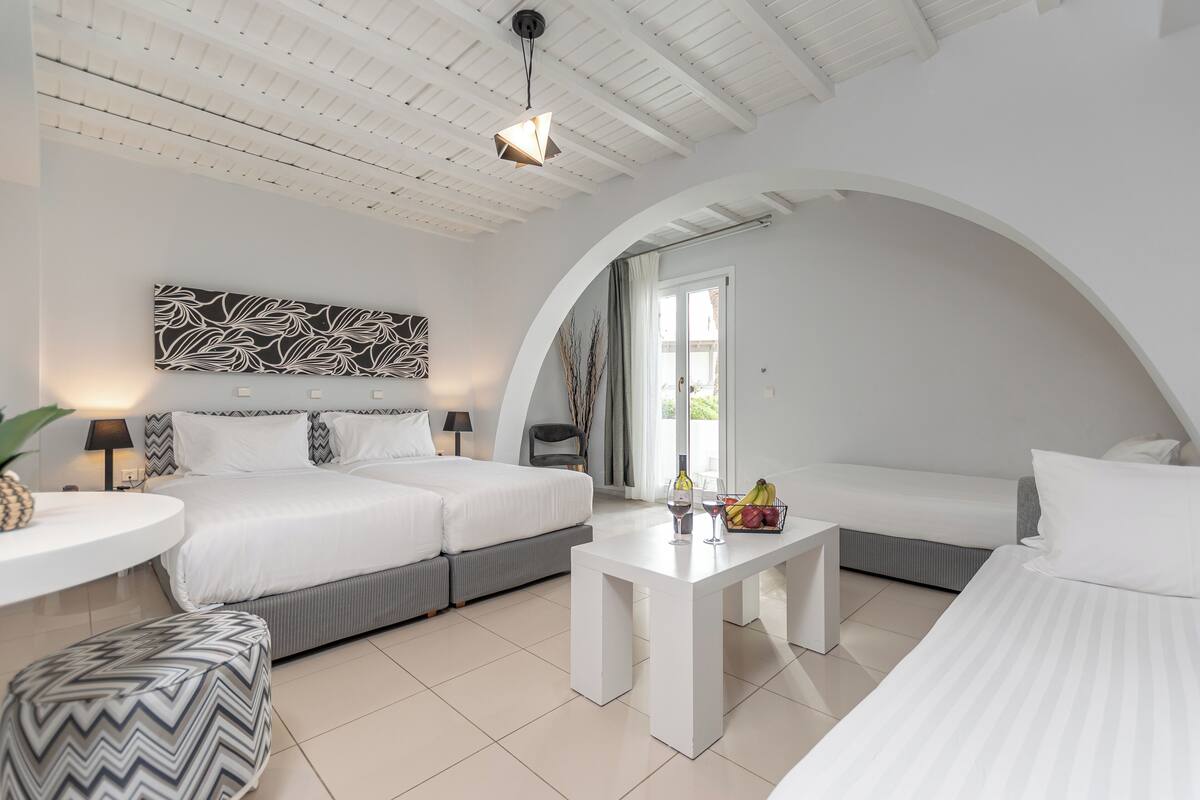 A modern hotel room with a large bed, a sofa, a dining table, and a ceiling with exposed beams.