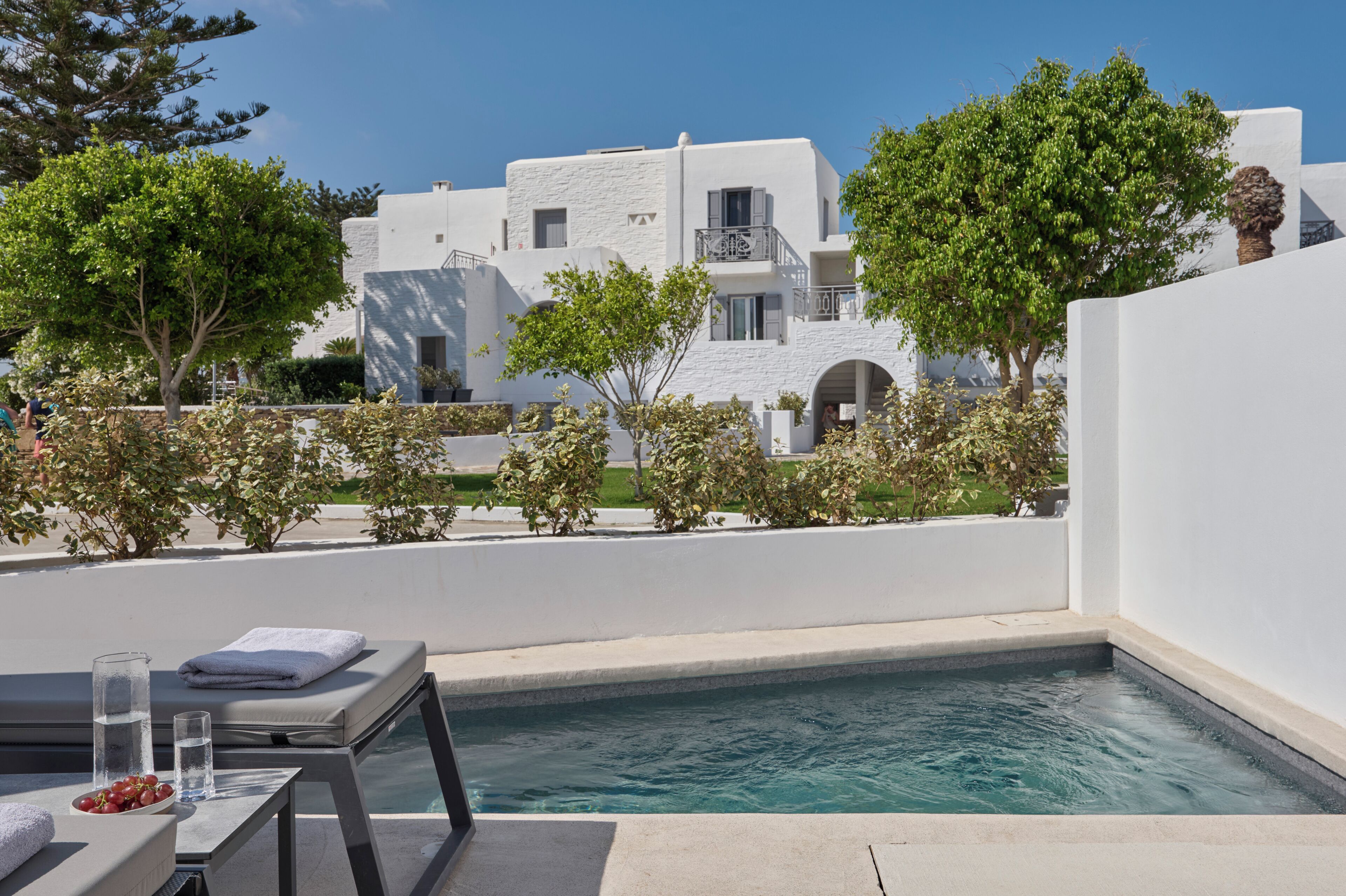 A pool area with a lounge chair, a table with a bowl of fruit, and a clear view of a white building with balconies.