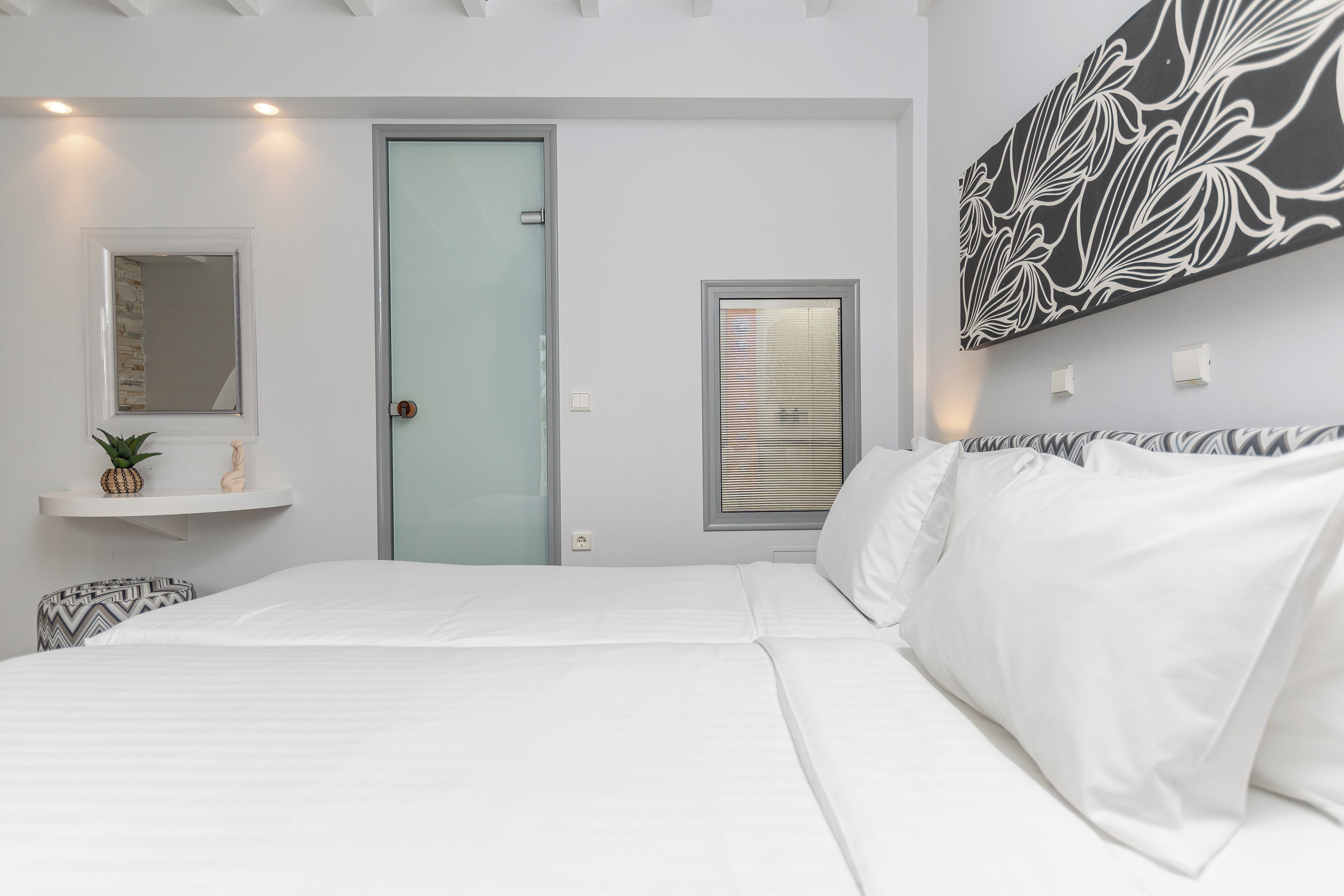 A bedroom with a large bed, a door, a window with blinds, a wall-mounted shelf with a pineapple and a small statue, and a wall with a black and white abstract painting.