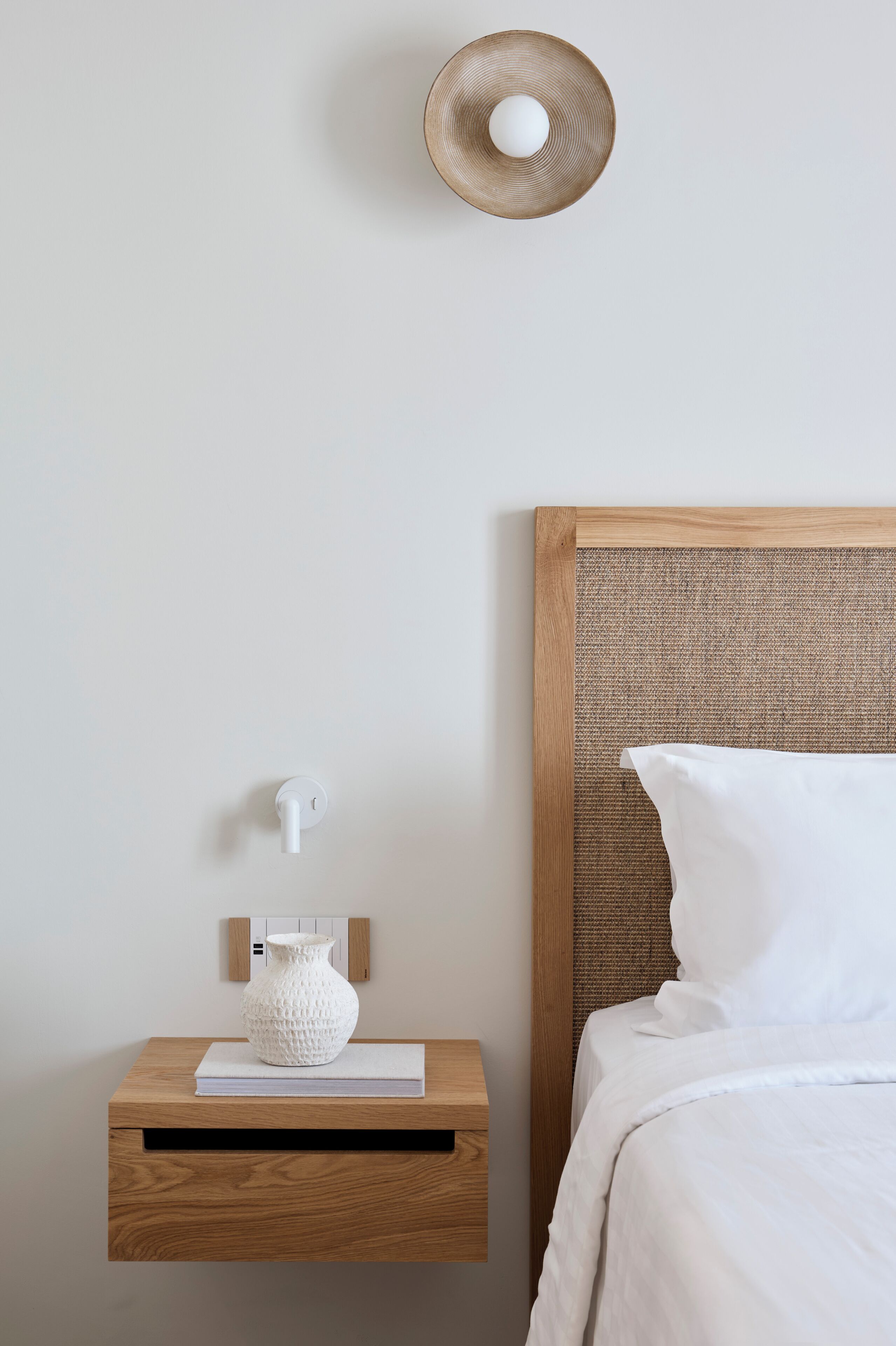 Habitación de hotel impecable con una mesita de noche de madera, un jarrón blanco, un libro y una cama con ropa blanca.