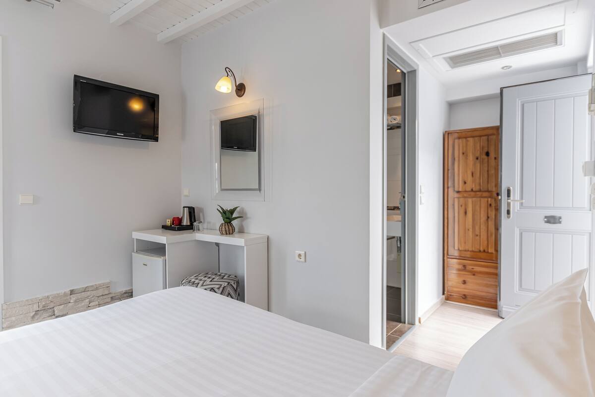 A modern bedroom with a bed, a TV mounted on the wall, a small desk with a plant, and a wooden door.
