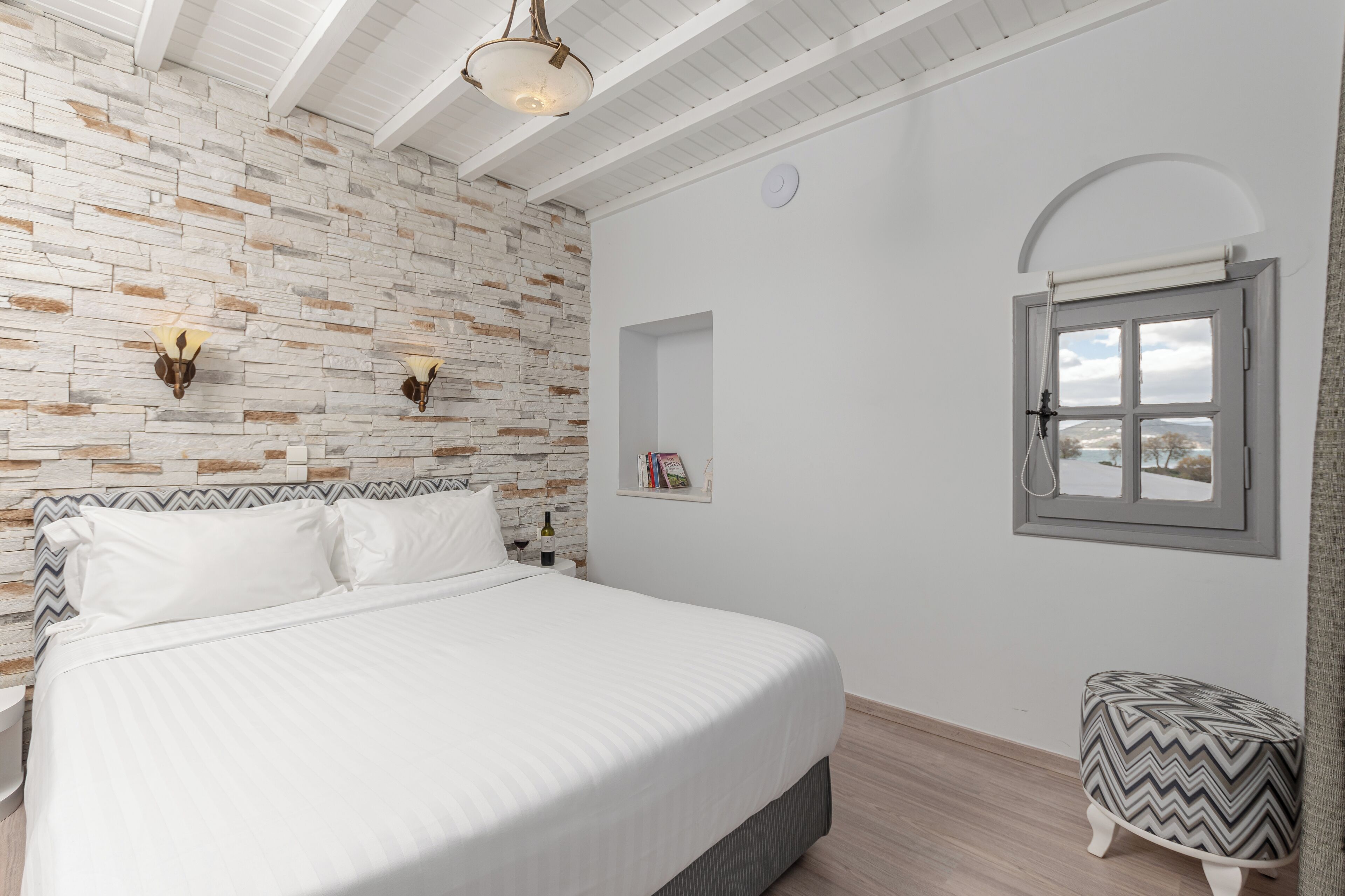 A bedroom with a brick-patterned wall, a bed with white bedding, a small bedside table with a wine bottle, and a window with a view.