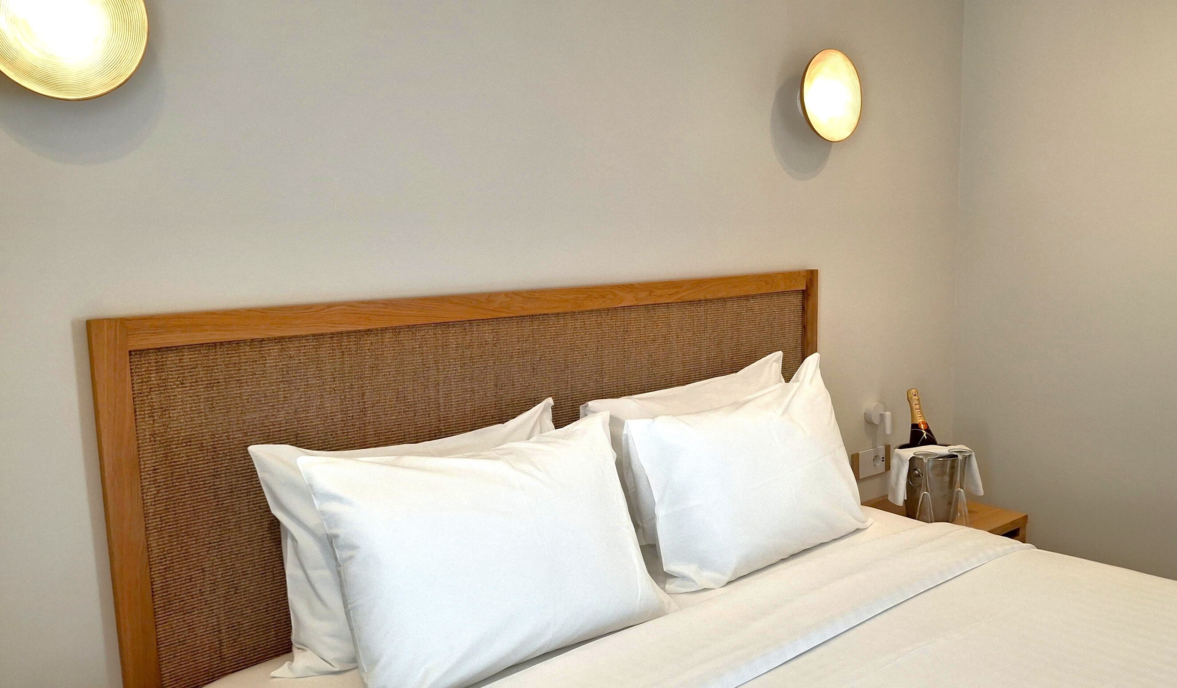Una cama bien hecha con sábanas blancas, un cabecero de madera y dos lámparas fijadas a la pared.