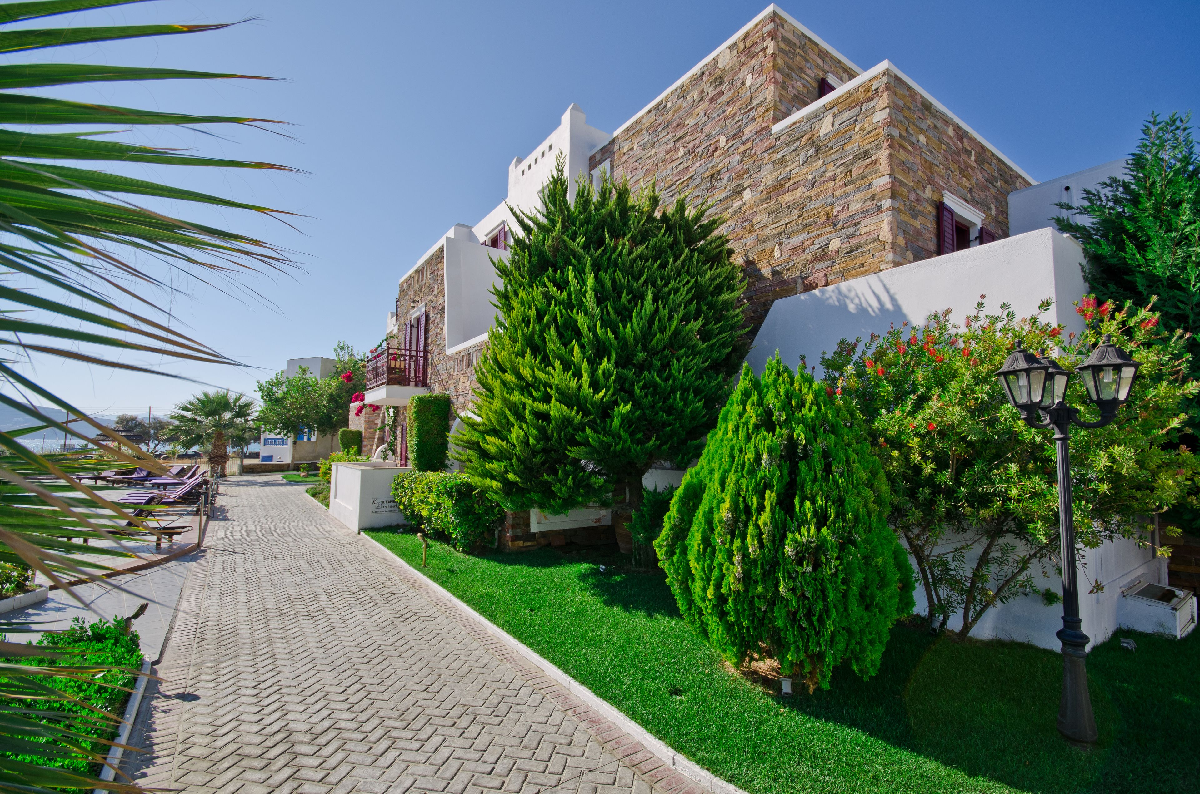A paved pathway leading to a building with stone and white walls, surrounded by well-maintained greenery and a clear sky.
