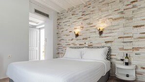 Un dormitorio con una cama grande, ropa de cama blanca, un aplique en la pared y una mesita de noche con una botella y un vaso.
