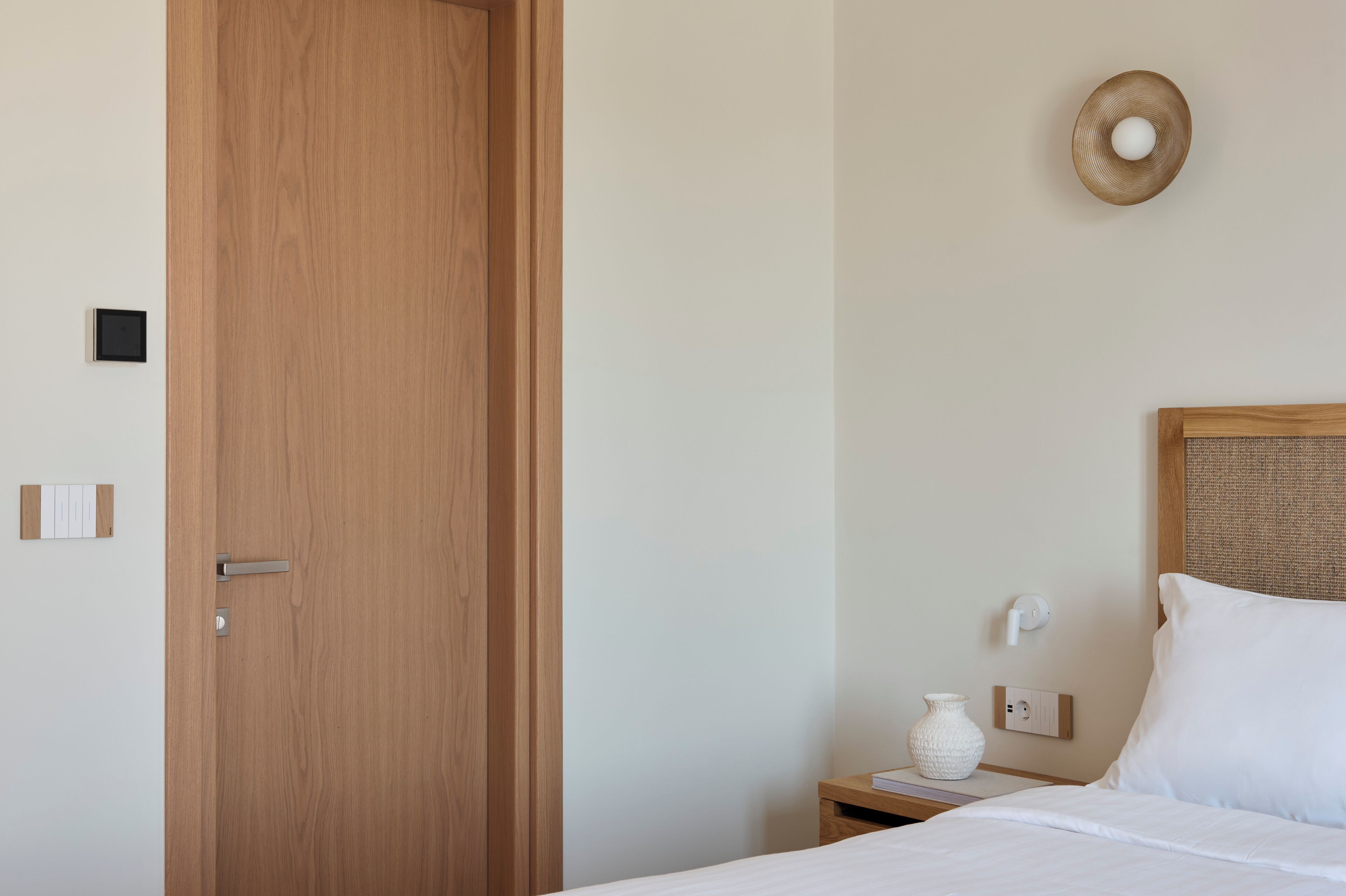 Habitación de hotel con puerta de madera, una mesita de noche con un florero, un enchufe de pared y una cama con ropa blanca.