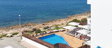 A pool area with a view of the sea, a white building with a sign, and a parked vehicle.