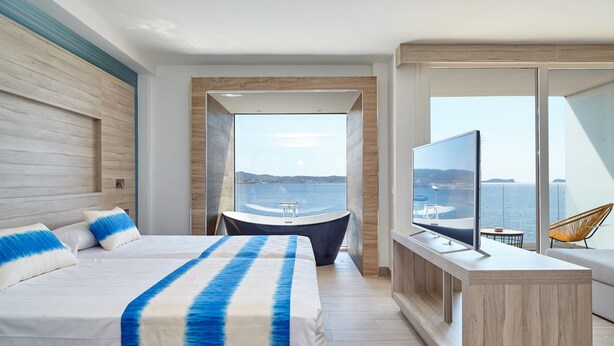 Ein modernes Hotelzimmer mit einem großen Bett, einer Badewanne, einem Fernseher und einem Balkon mit Meerblick.