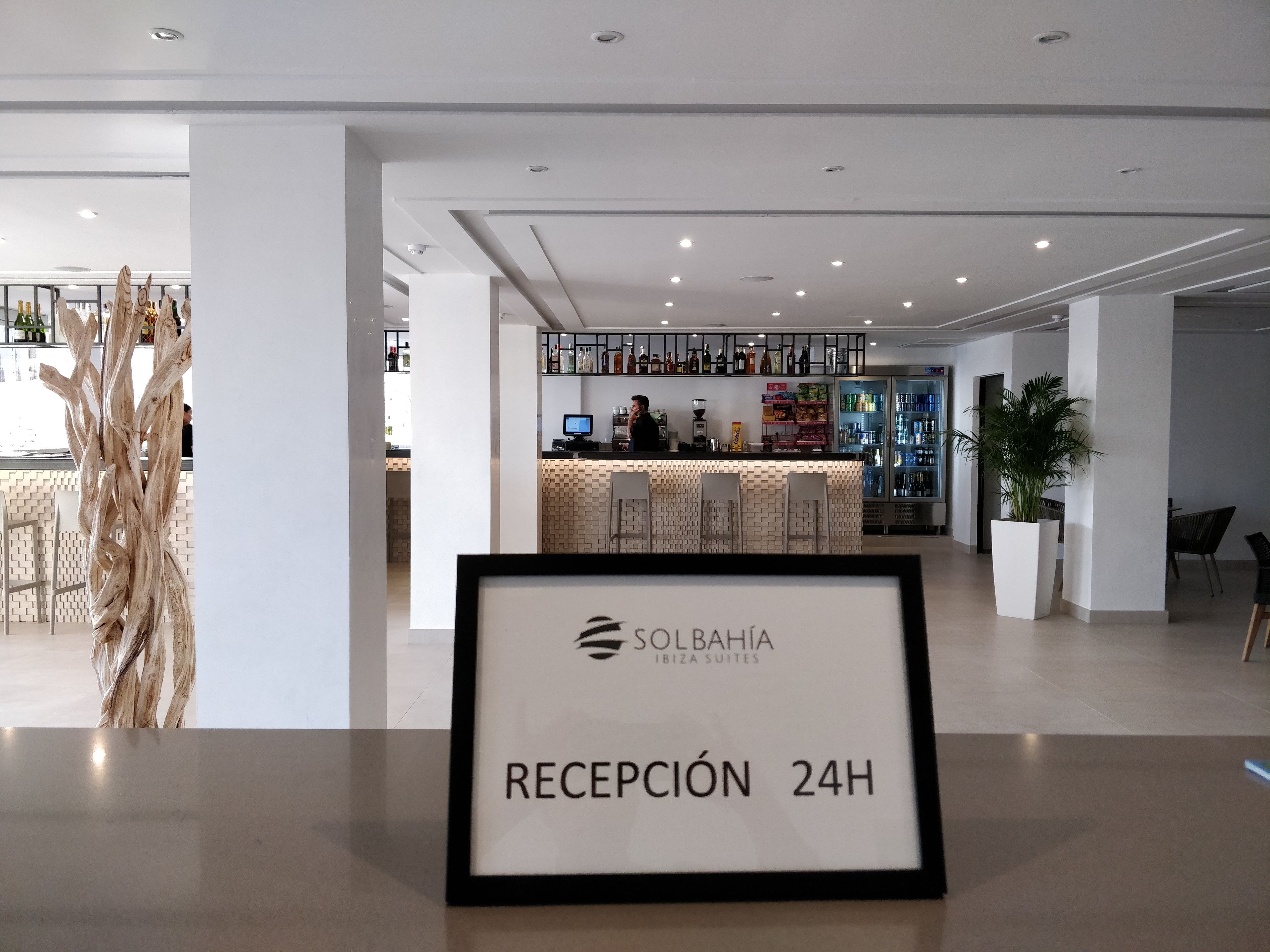 A reception area with a sign indicating 24-hour reception, a bar area with shelves of bottles, and a potted plant.