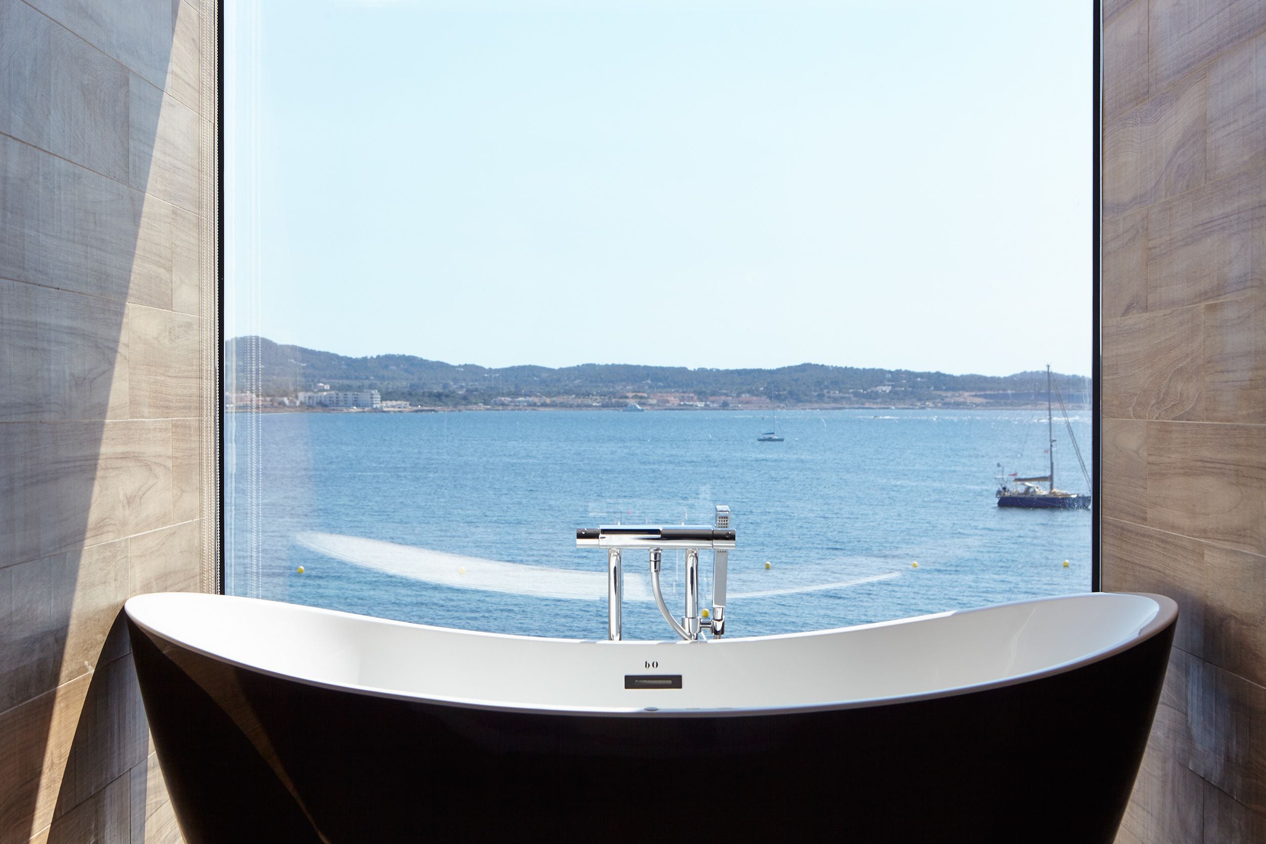 A modern bathroom with a freestanding bathtub, a view of the sea, and a sailboat in the distance.