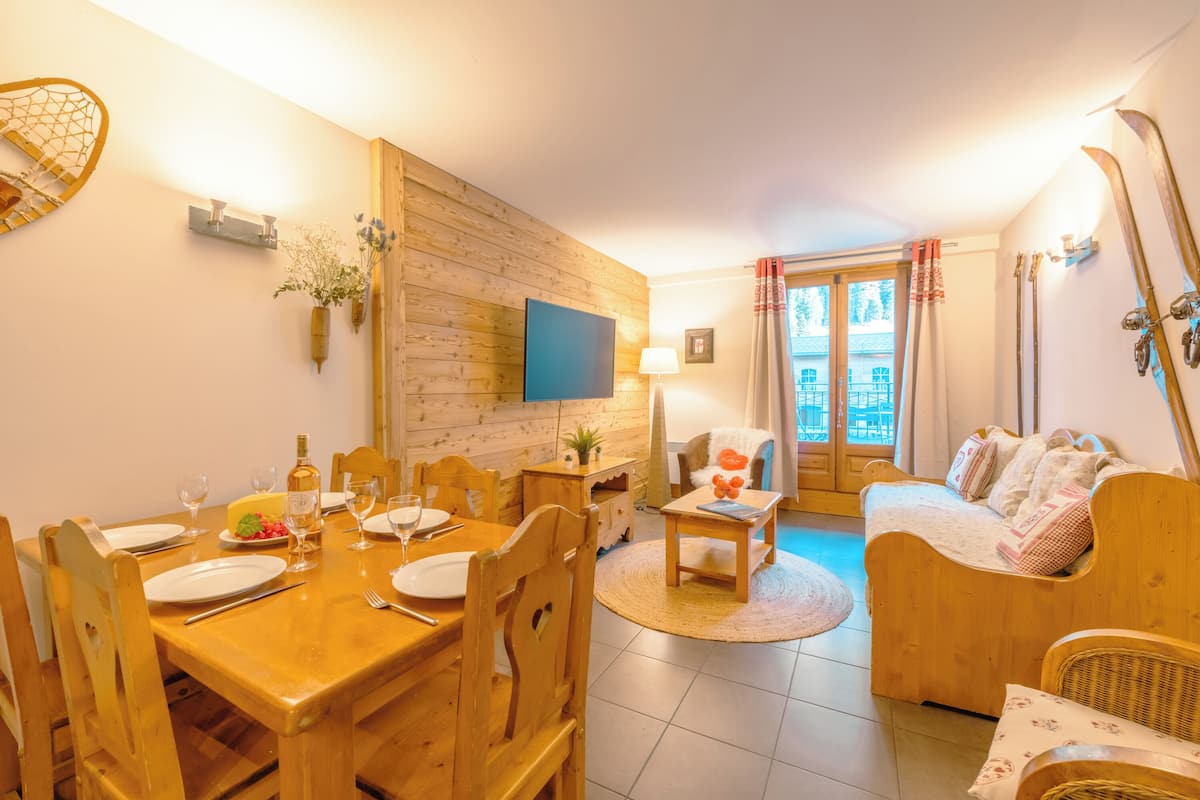 Un salon chaleureux avec une table à manger en bois, préparée pour quatre personnes, un canapé et un téléviseur fixé au mur.
