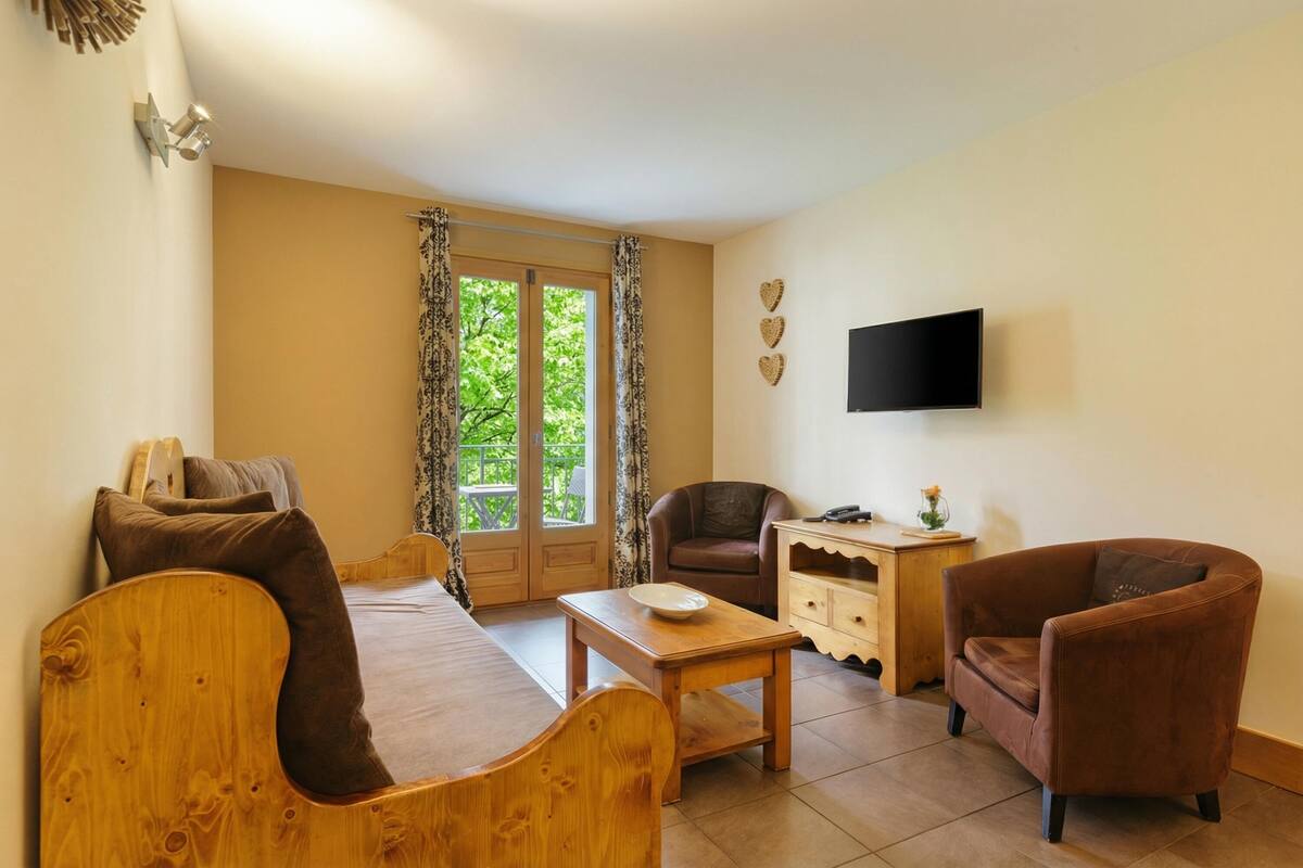 Une pièce avec un lit en bois, une table en bois, deux fauteuils, un téléviseur et une fenêtre avec des rideaux.